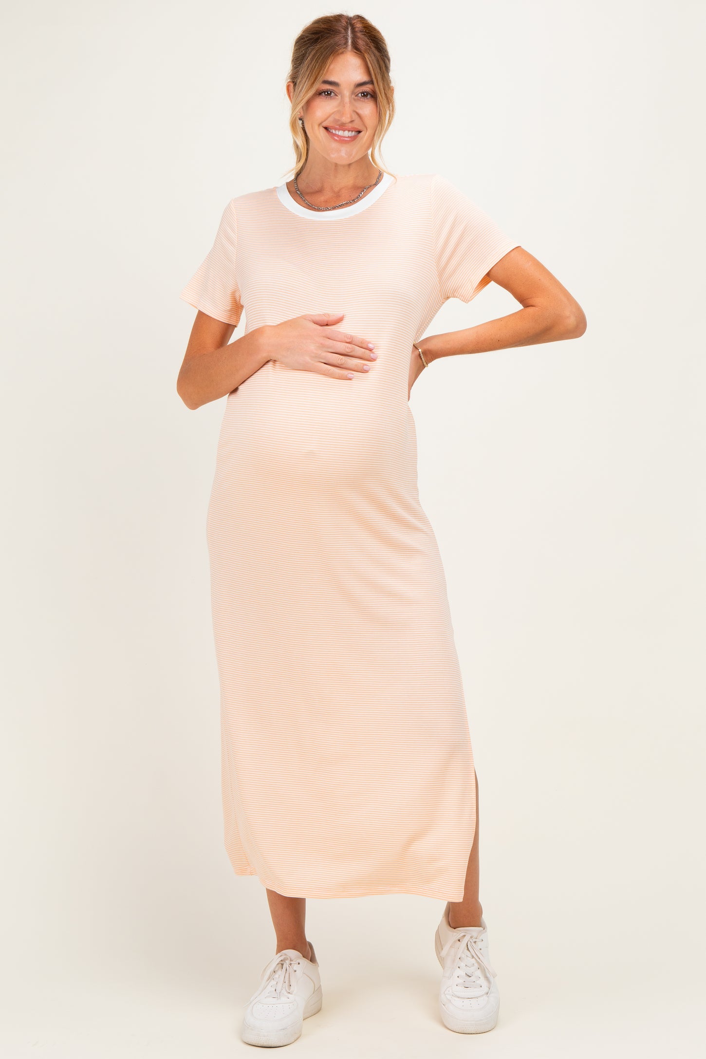 woman wearing a light peach maternity maxi dress with short sleeves and a white crew neckline, holding her baby bump
