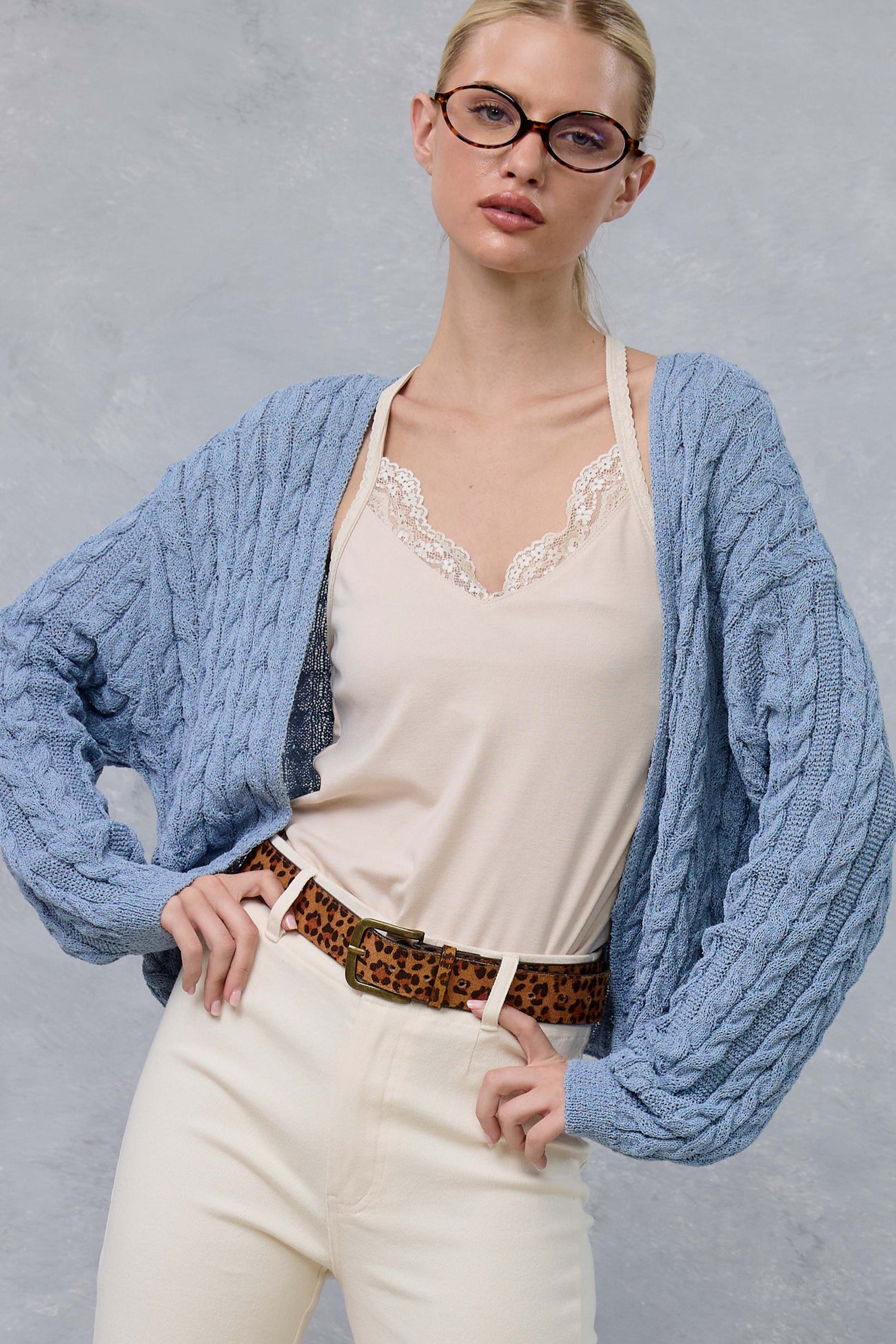 woman wearing a light blue cable knit cardigan over a cream lace camisole and high-waisted cream pants with a leopard print belt