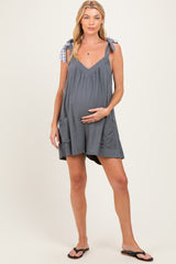 woman wearing a gray maternity dress with plaid tie straps and pockets, gently holding her baby bump