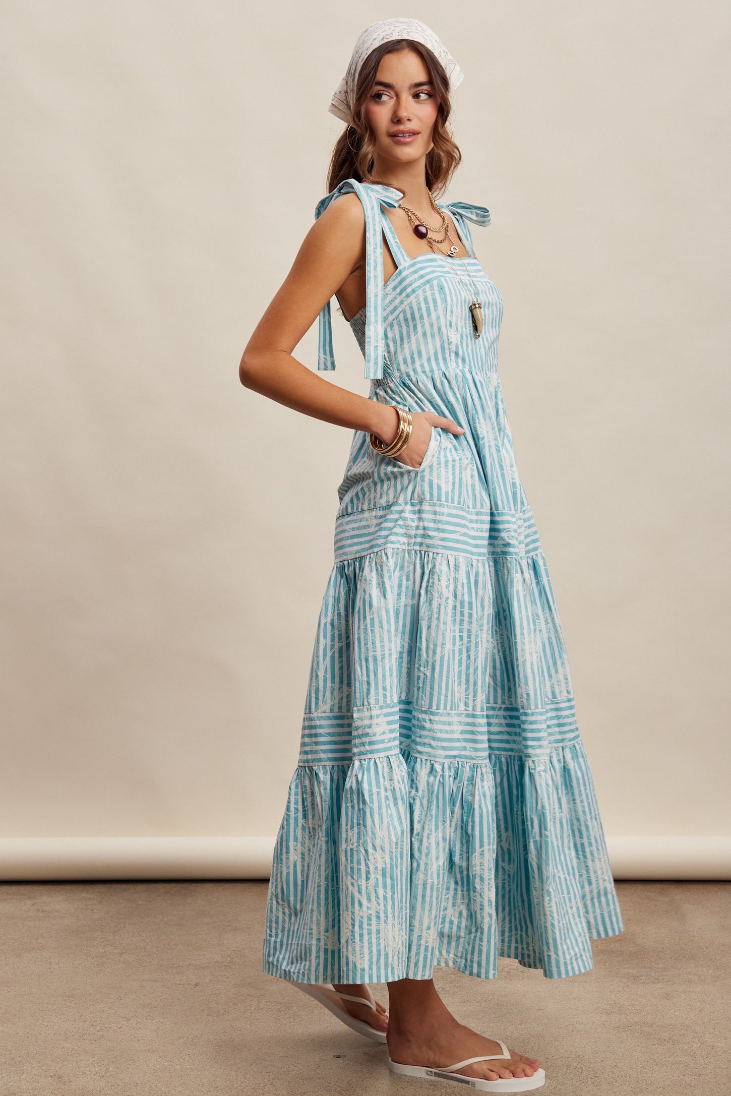 woman wearing a light blue striped maxi dress with tie straps and pockets, standing sideways with a headscarf and sandals