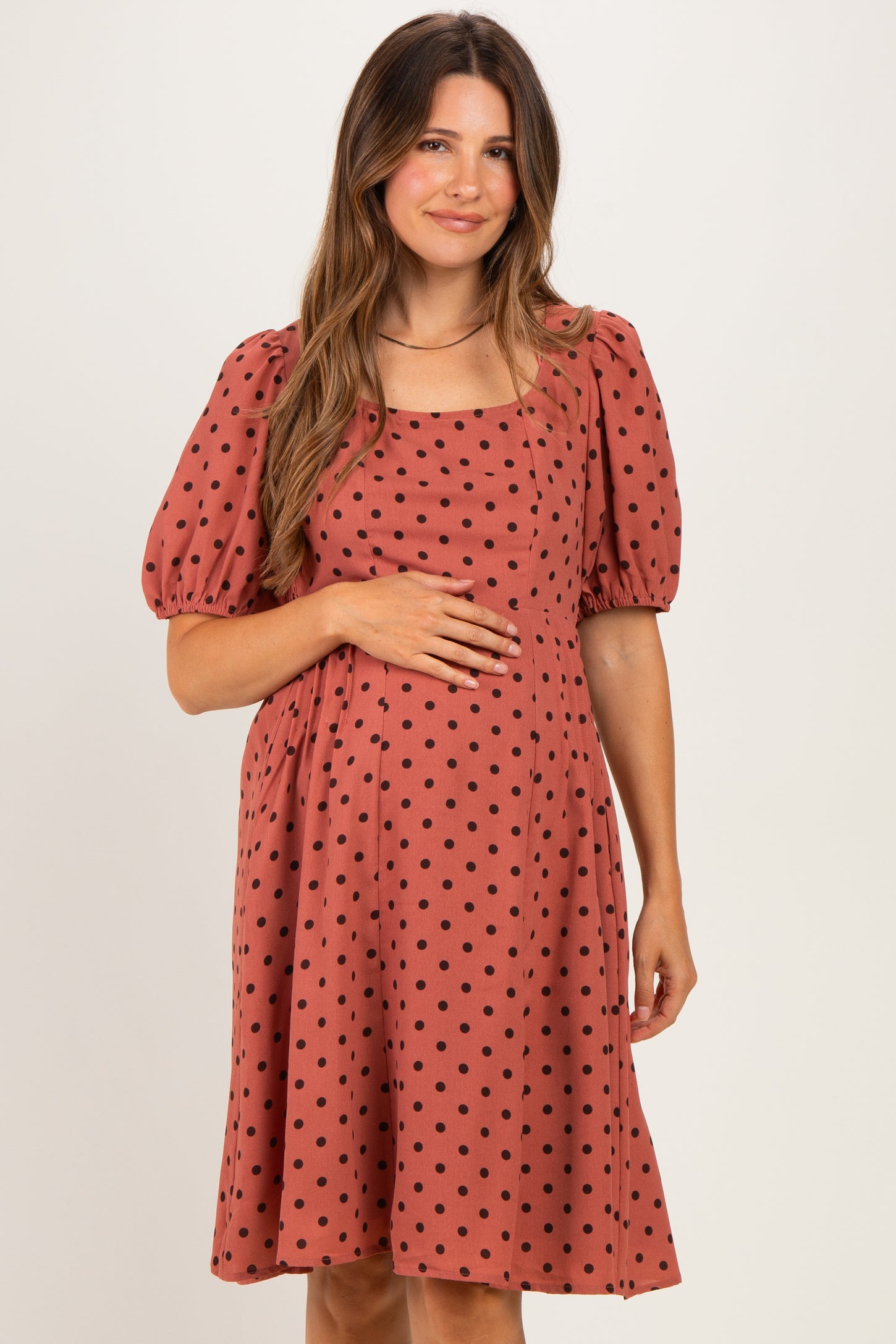 woman wearing a rust colored maternity dress with puff sleeves and black polka dots, holding her baby bump