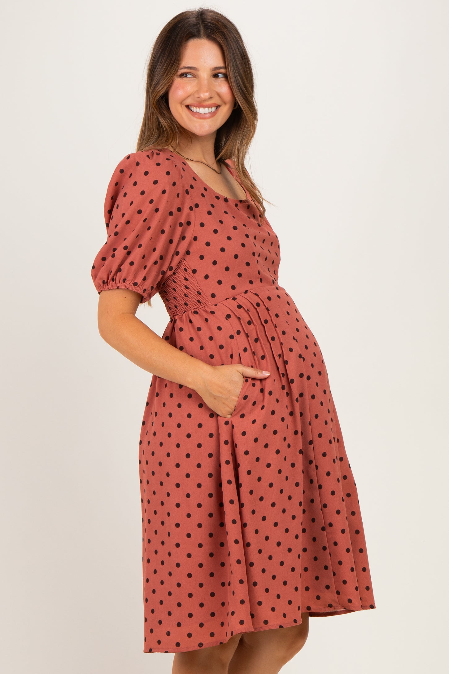 woman wearing a rust polka dot maternity dress with a cinched waist and pockets, showing her baby bump