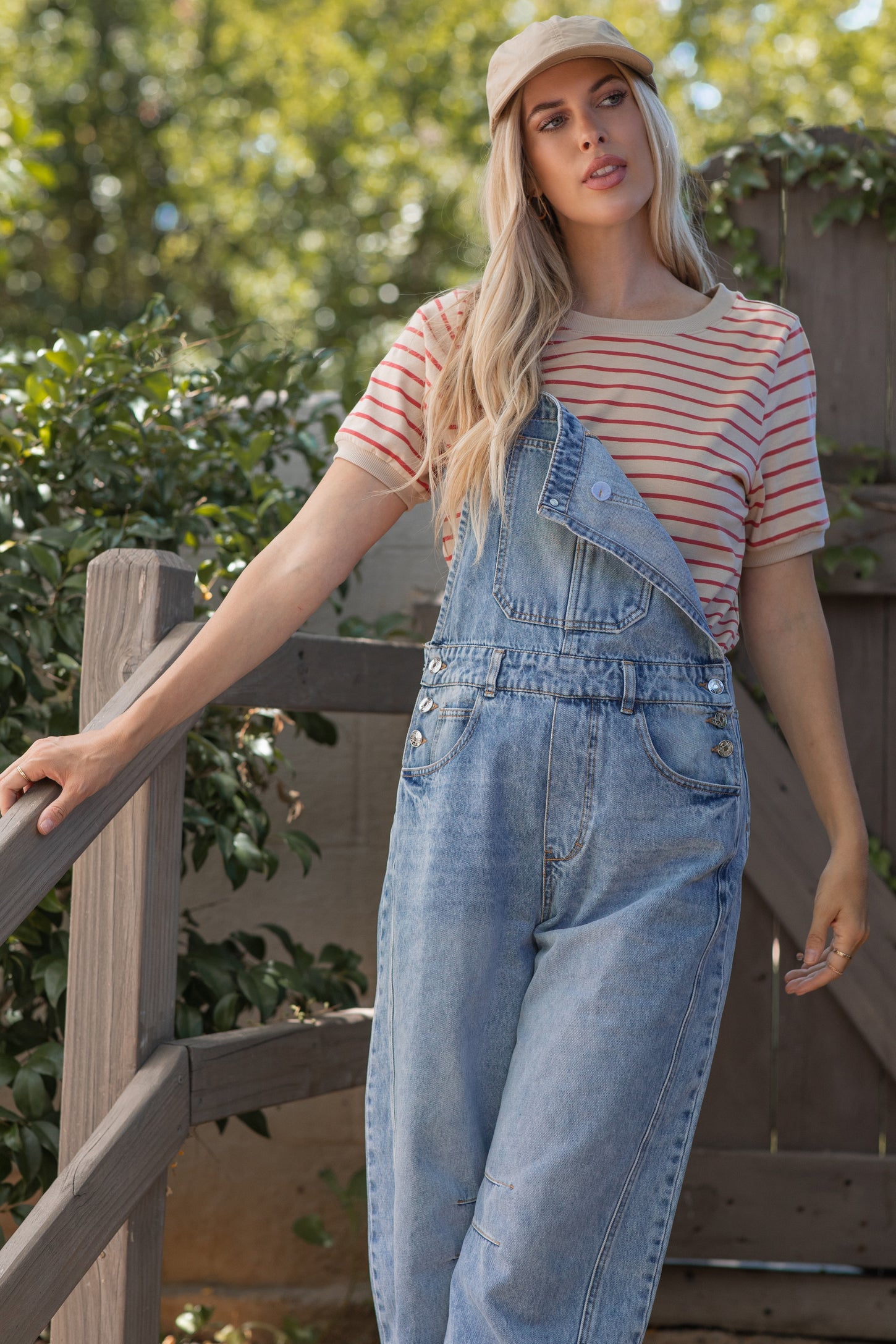 woman wearing a striped short sleeve t-shirt and blue denim overalls, standing outdoors with a cap on her head