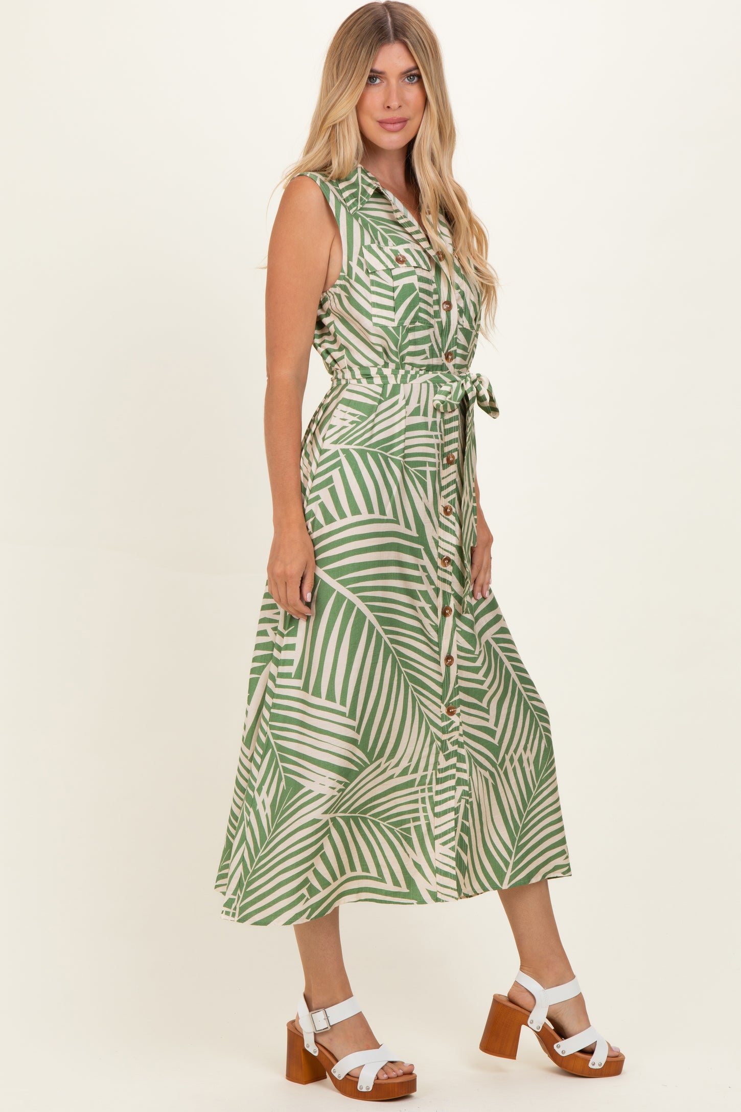 woman wearing a green and white palm print midi dress with a belted waist and buttons, standing on a neutral background
