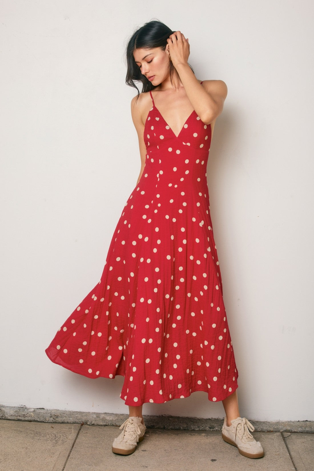 woman wearing a red polka dot summer dress with a v-neck and spaghetti straps, posing with one hand in her hair