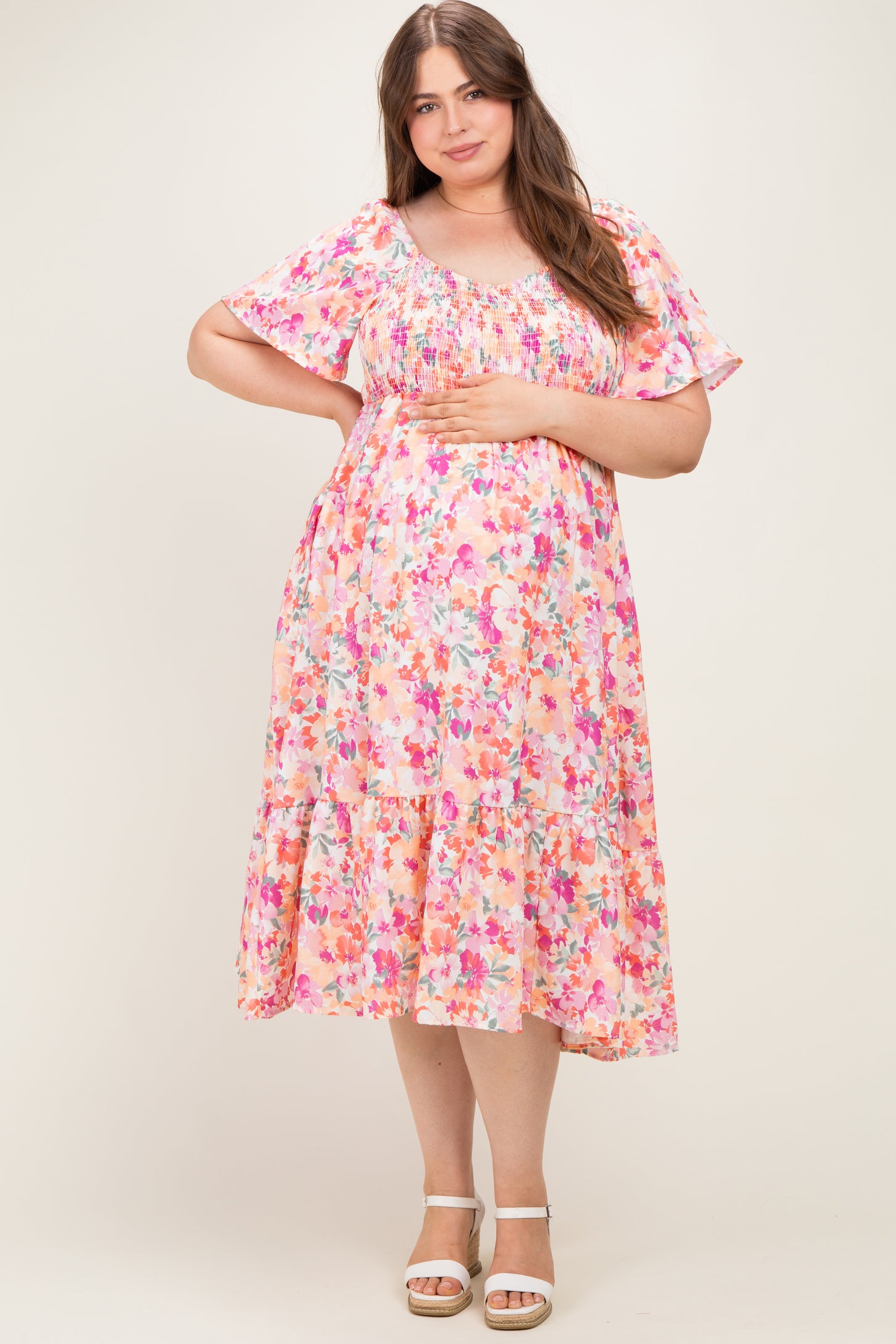woman wearing a floral pink maternity dress with short sleeves and a tiered skirt, gently holding her baby bump