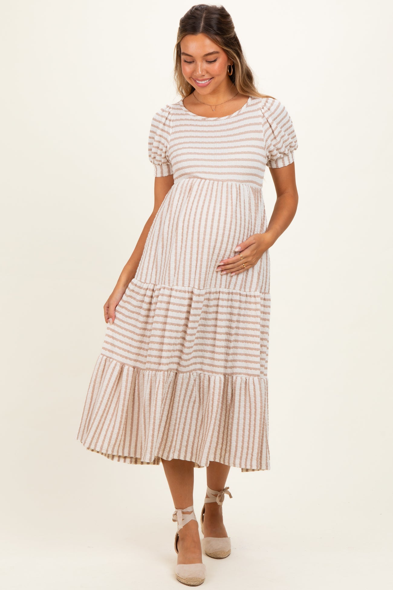 woman wearing a beige and striped maternity maxi dress with puff sleeves and a ruffled hem, holding her baby bump