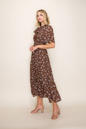woman wearing a brown floral midi dress with a ruffled neckline and short sleeves, standing against a neutral background