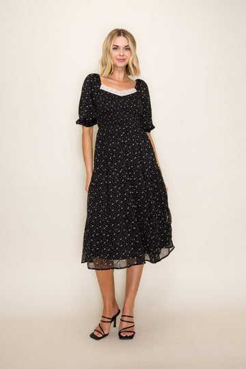 woman wearing a black floral midi dress with short puff sleeves and a square neckline, standing against a neutral background