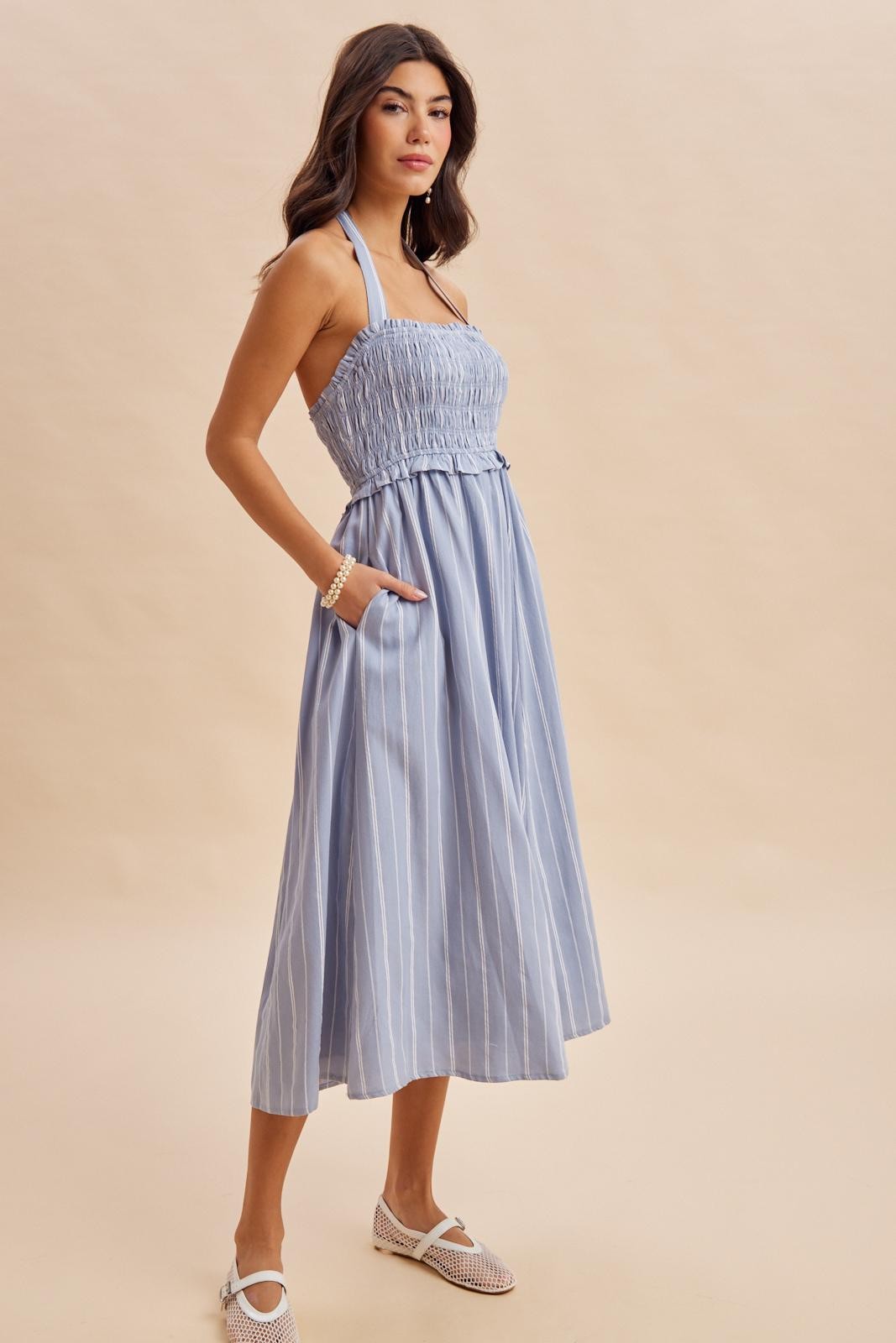 woman wearing a light blue striped midi dress with a smocked bodice and pockets, styled with a halter neckline