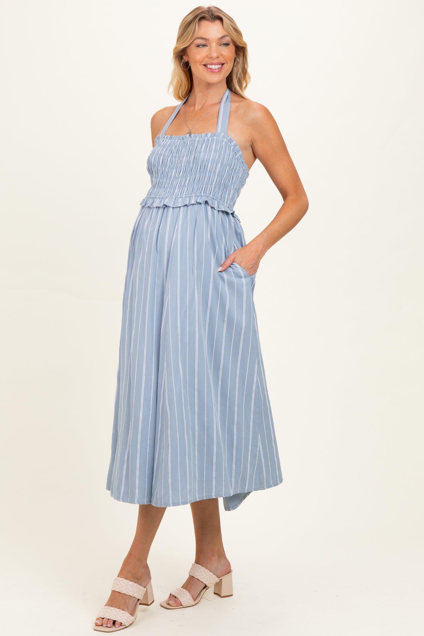woman wearing a light blue striped maternity dress with smocked bodice and pockets, smiling and showing her baby bump