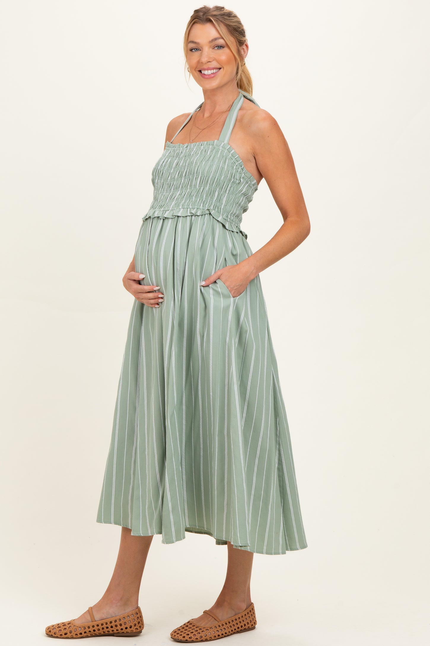 woman wearing a light green striped maternity dress with smocked bodice and pockets, holding her baby bump