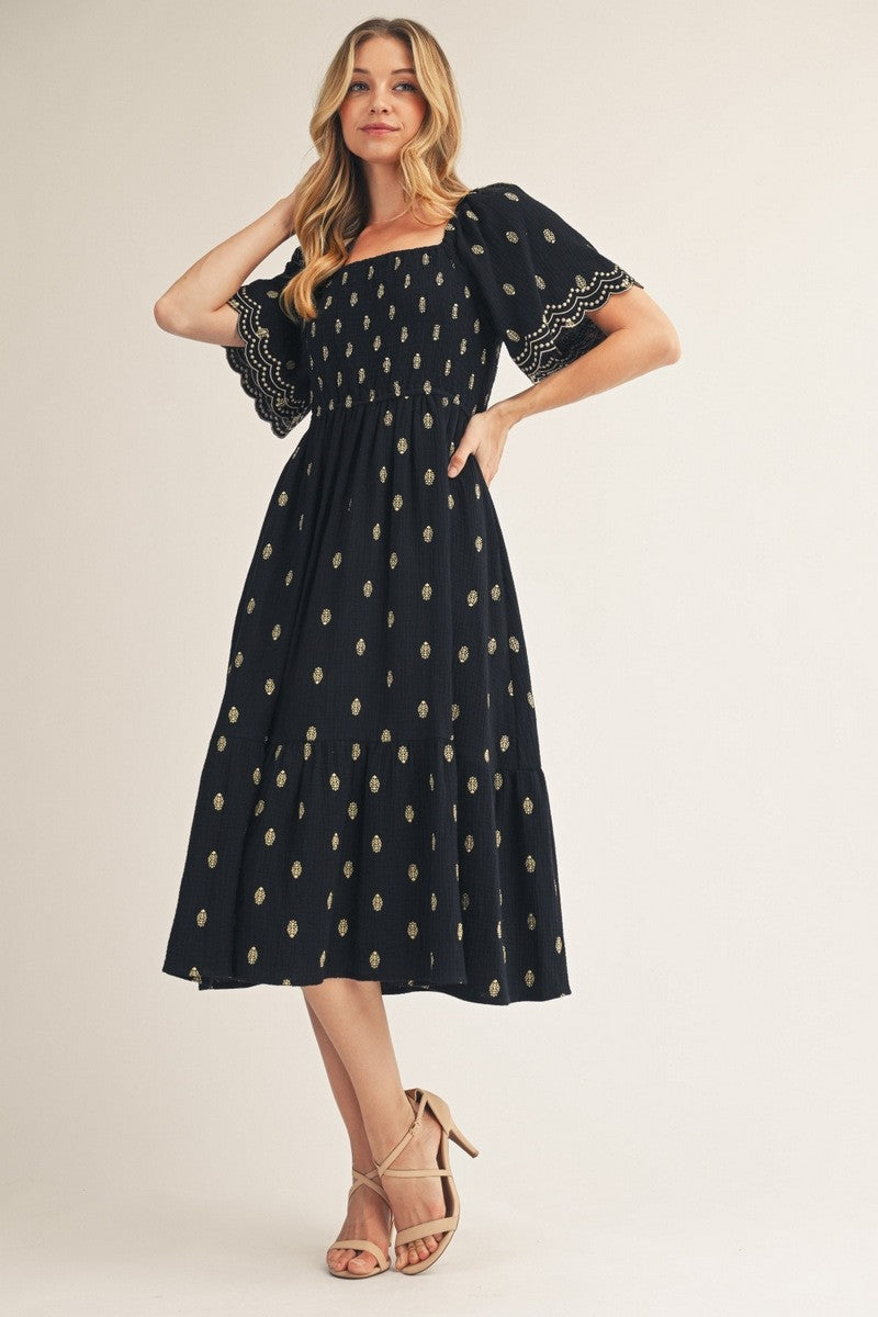 woman wearing a black midi dress with gold embellishments and puff sleeves, standing elegantly with one hand on her hip