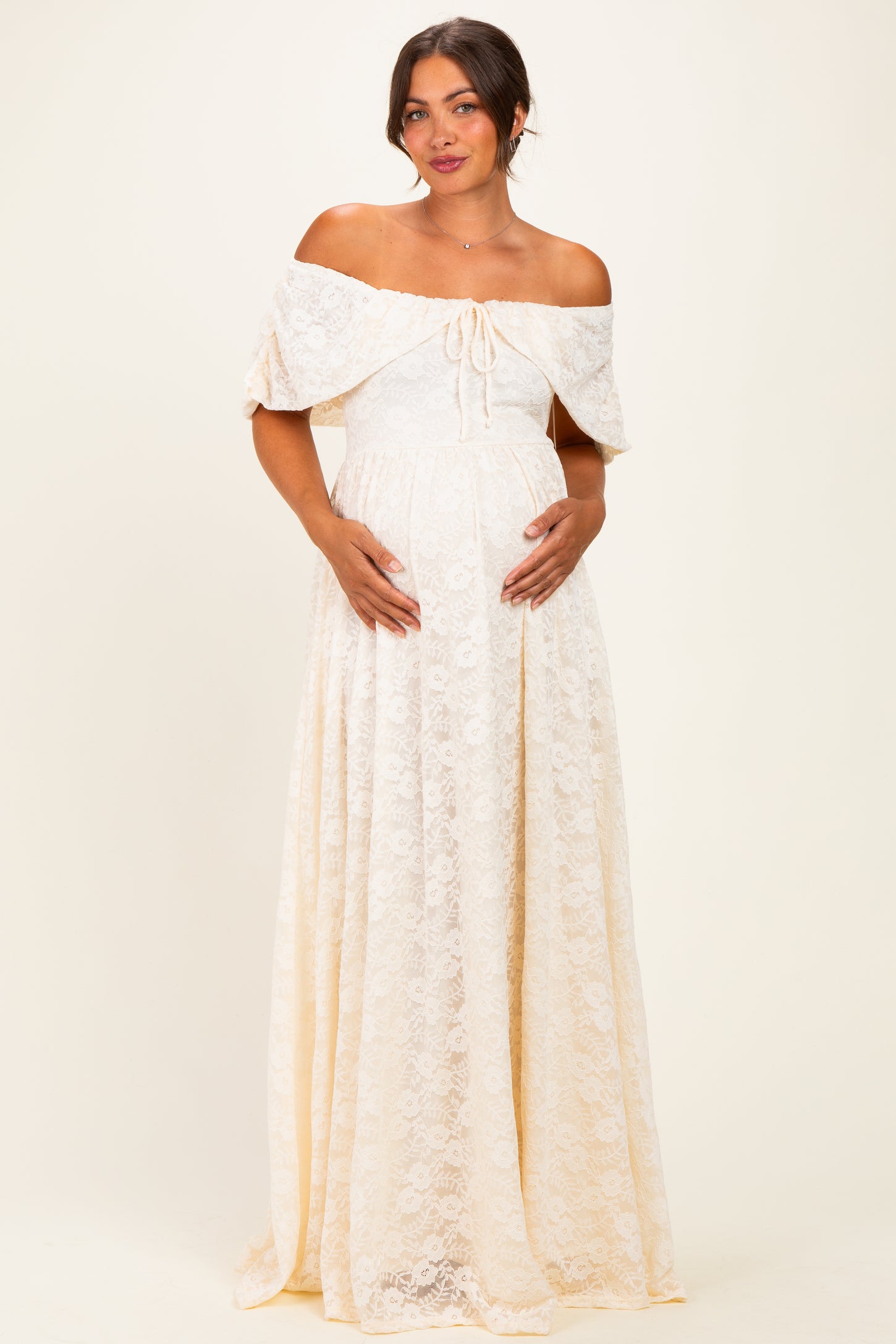 woman wearing a cream off-the-shoulder lace maternity maxi dress and cradling her baby bump