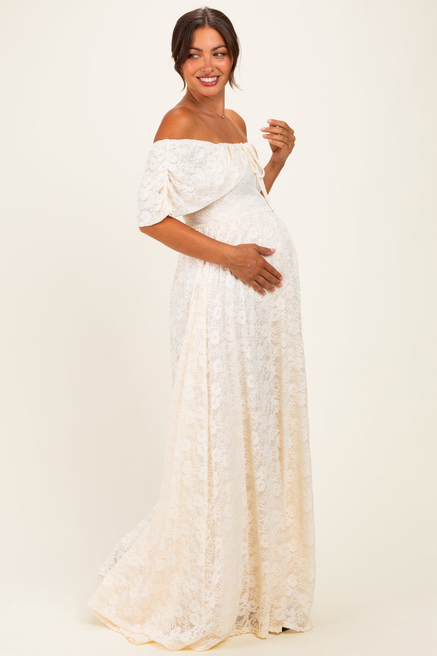 woman wearing an off-the-shoulder cream lace maternity dress and gently cradling her baby bump