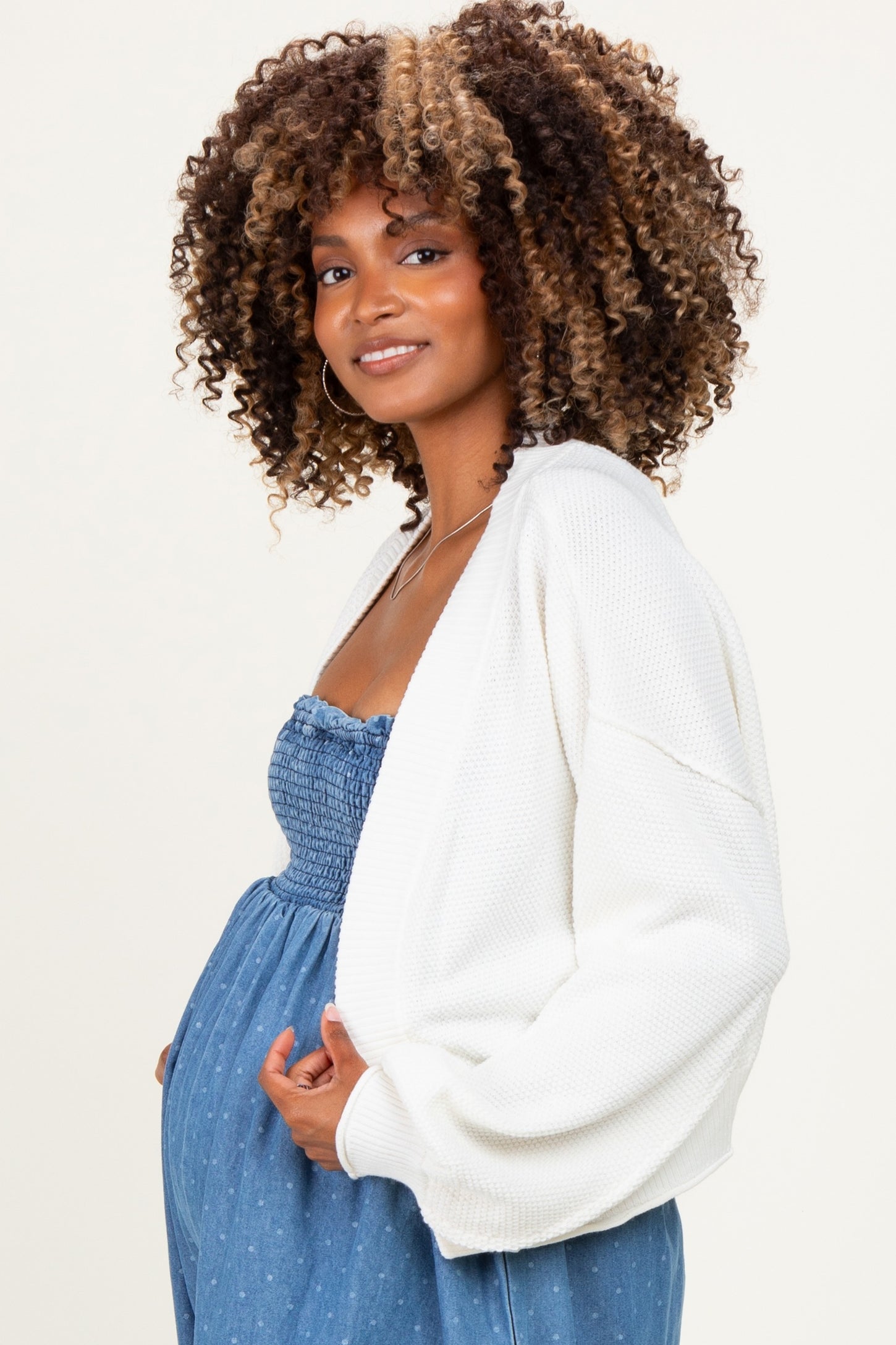 woman wearing a white knit maternity cardigan over a blue smocked maternity dress with a visible baby bump
