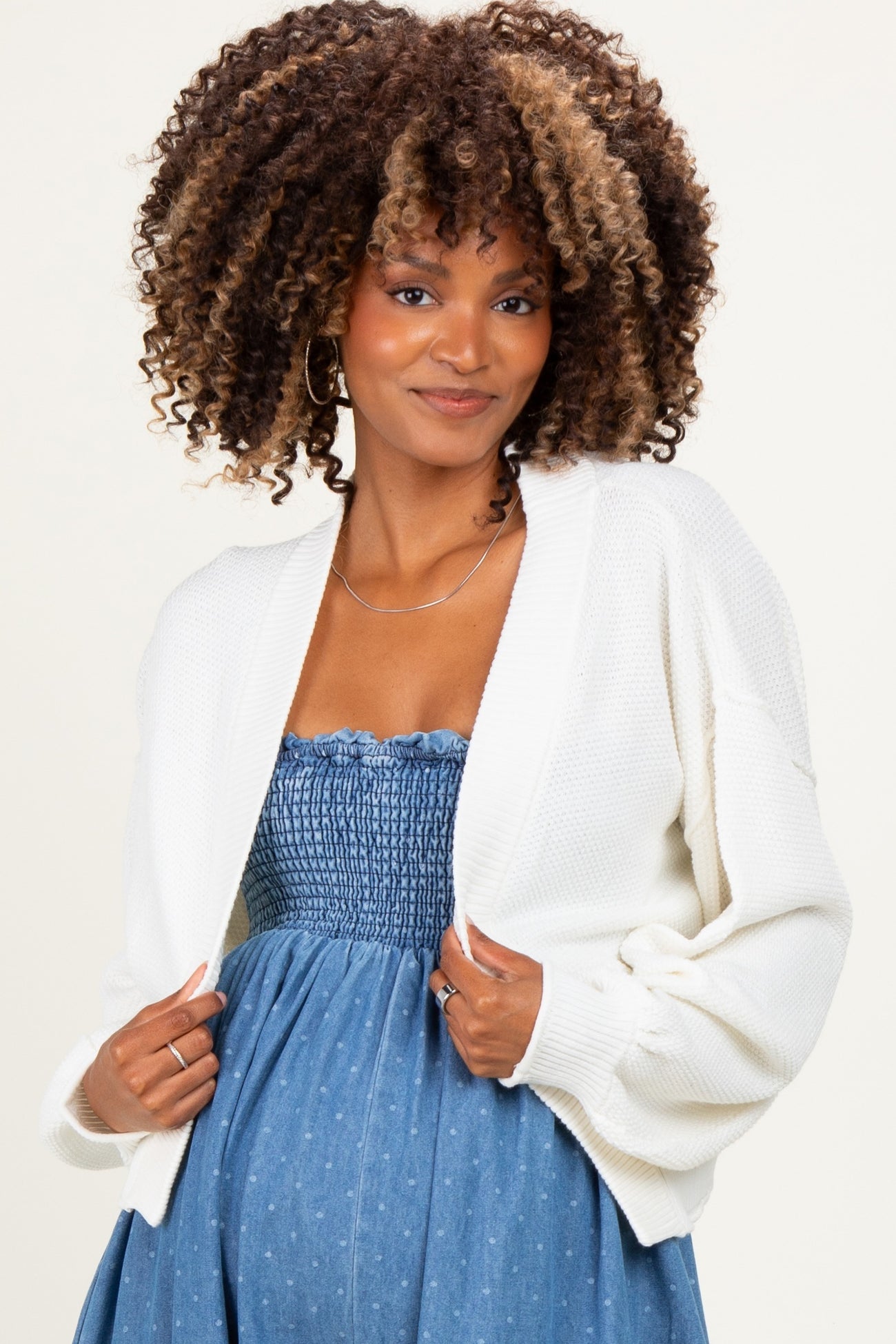 woman wearing a white cardigan over a blue smocked maternity dress and showing her baby bump