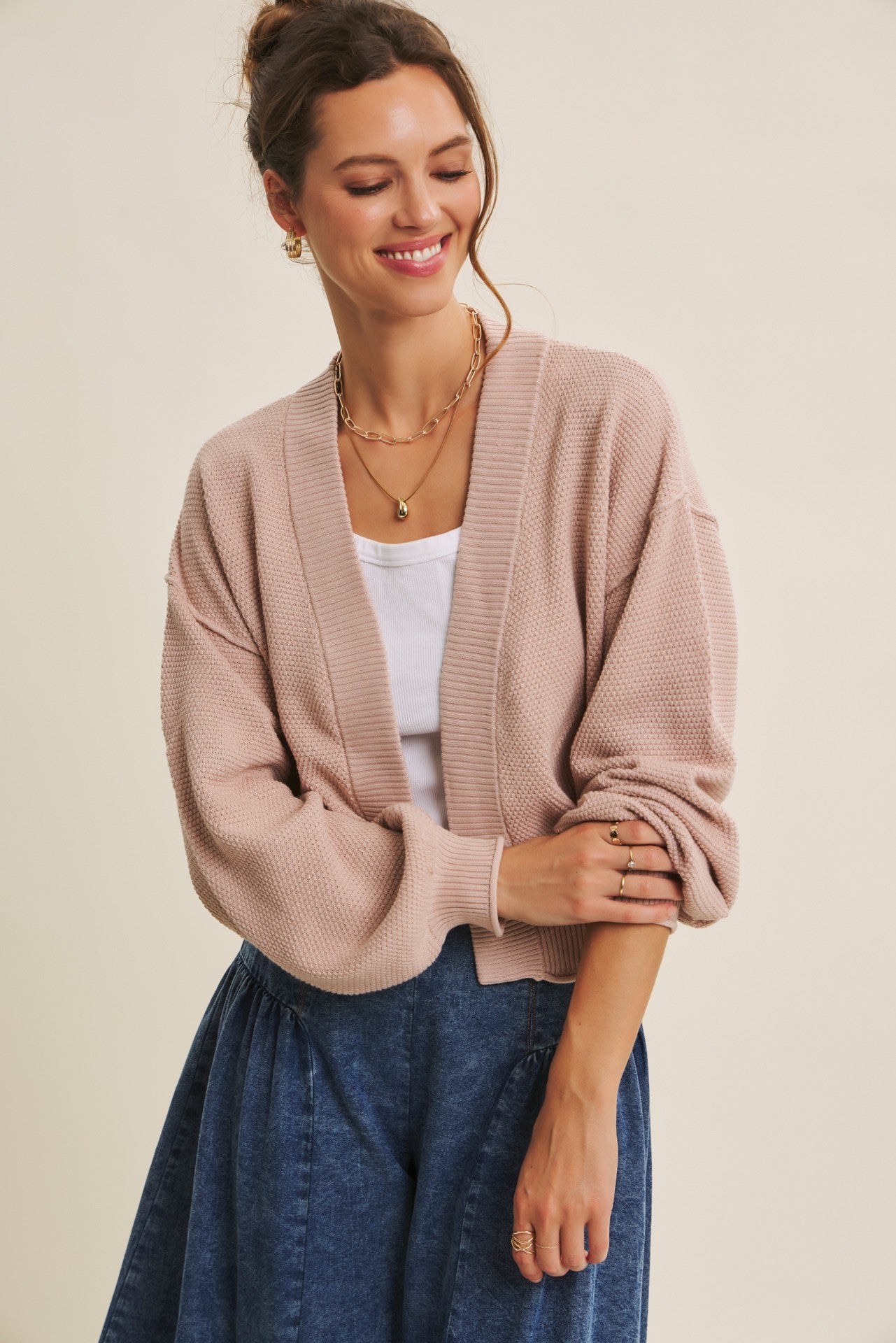 woman wearing a light pink oversized cardigan over a white top and a denim skirt, smiling and standing with one hand on her hip