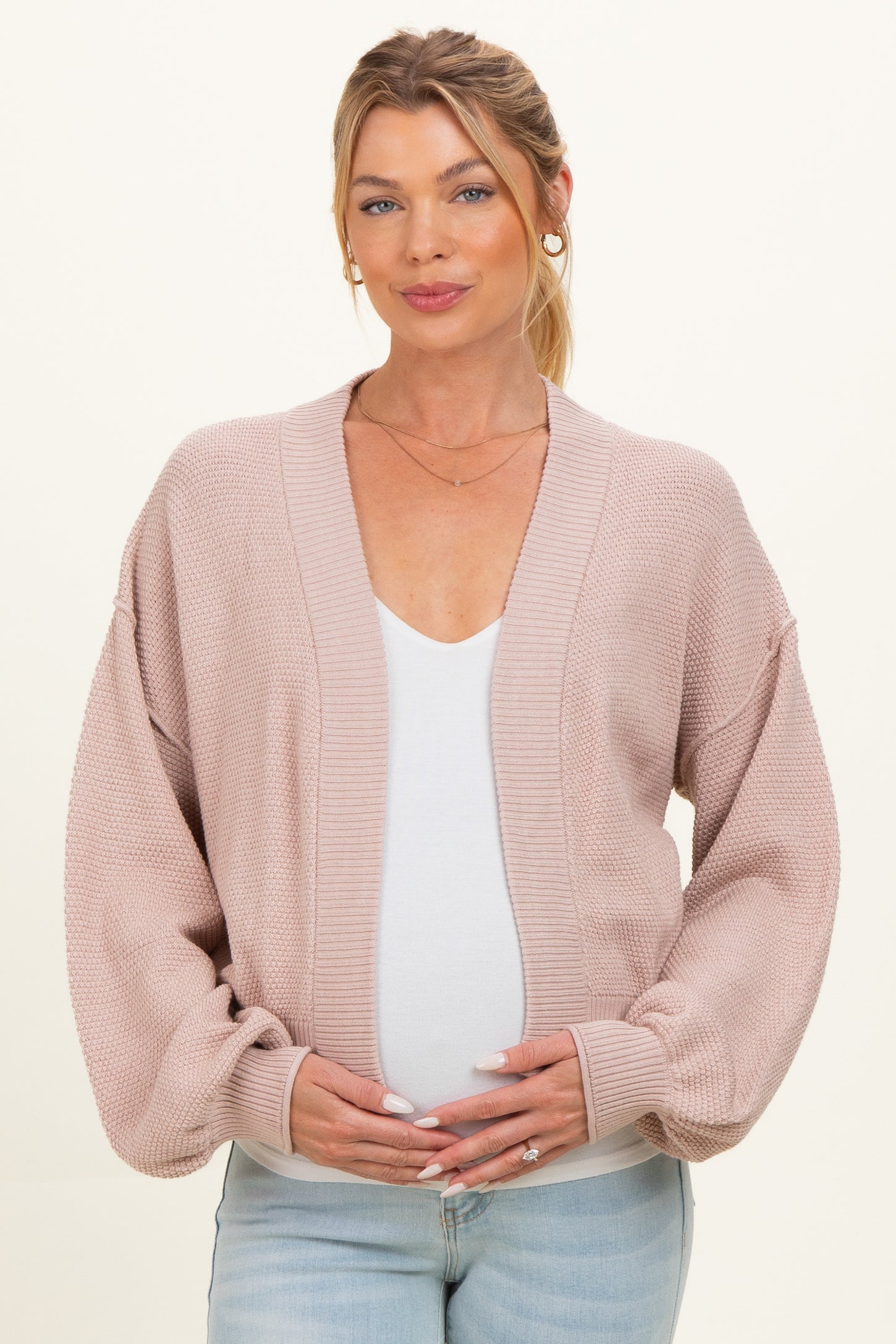 woman wearing a light pink maternity cardigan over a white top and holding her baby bump