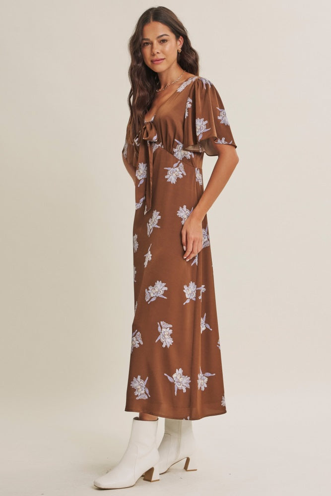 woman wearing a brown floral maxi dress with short sleeves and a tie detail at the waist, paired with white ankle boots