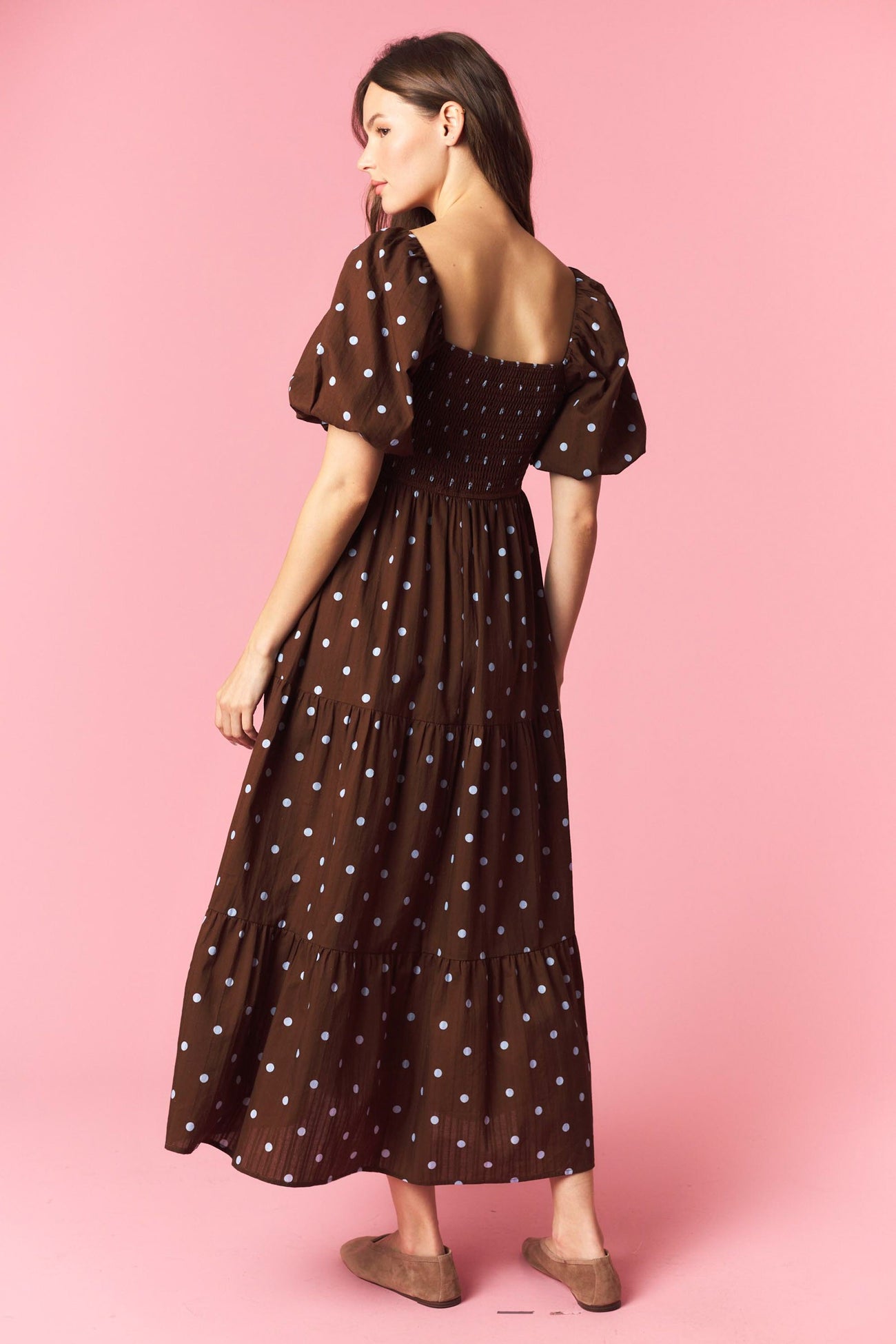 woman wearing a brown polka dot maxi dress with puff sleeves and smocked back, standing against a pink background