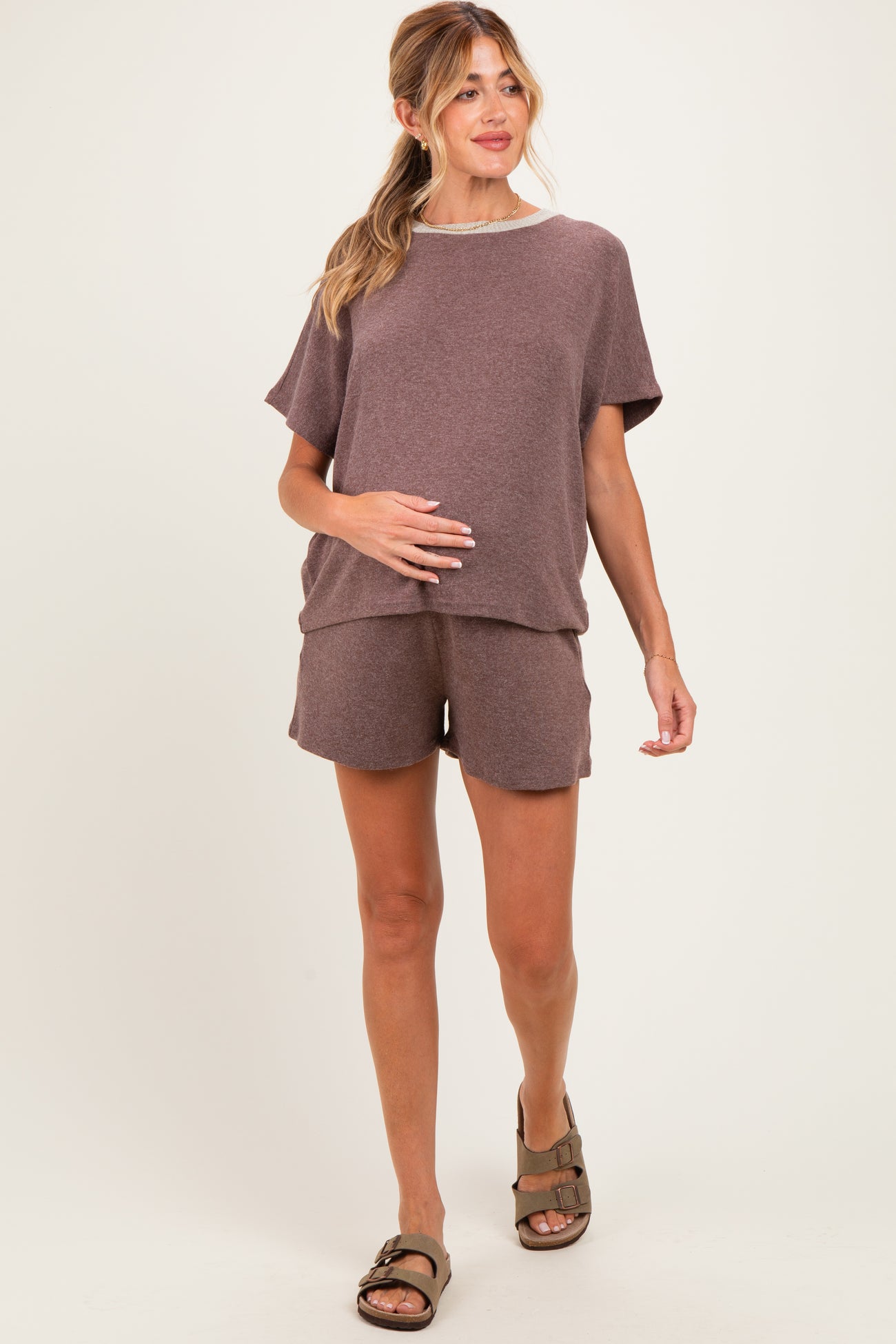 woman wearing a brown maternity loungewear set with a loose top and shorts, holding her baby bump