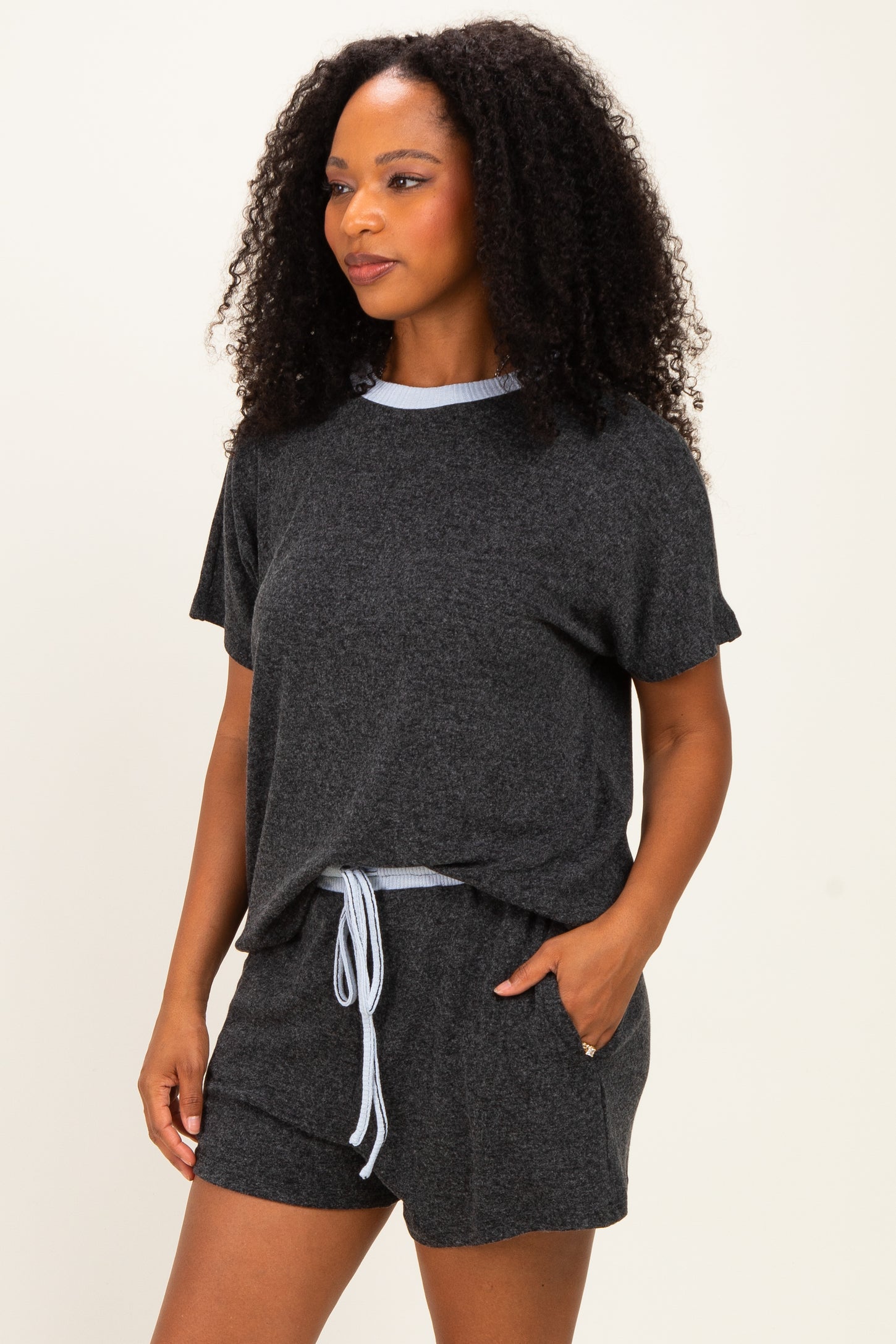 woman wearing a dark gray short sleeve lounge set with a light blue trim and drawstring shorts, posing with one hand in pocket