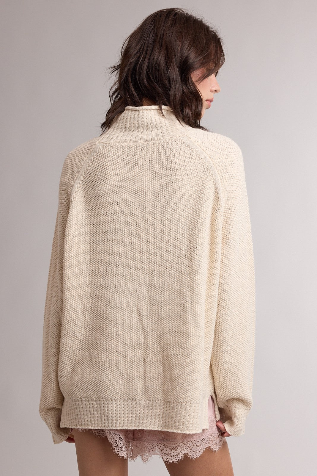 woman wearing a cream oversized knit sweater with a high neck and ribbed hem, shown from the back