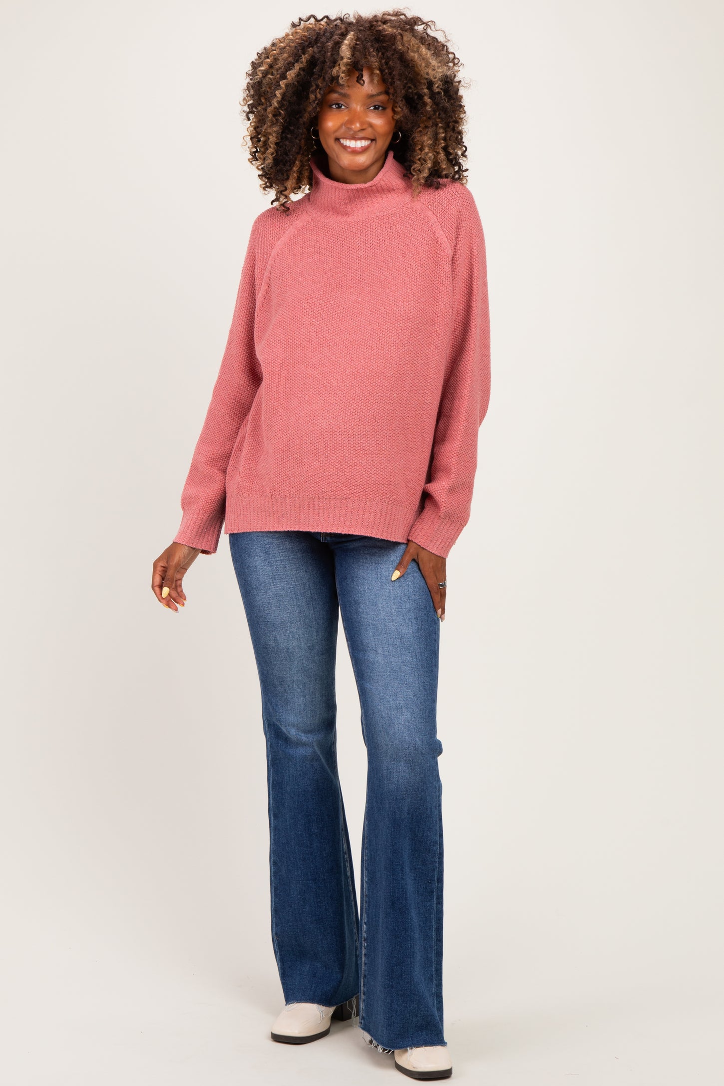 woman wearing a pink maternity sweater with a high neckline and flared jeans, smiling and showing her baby bump