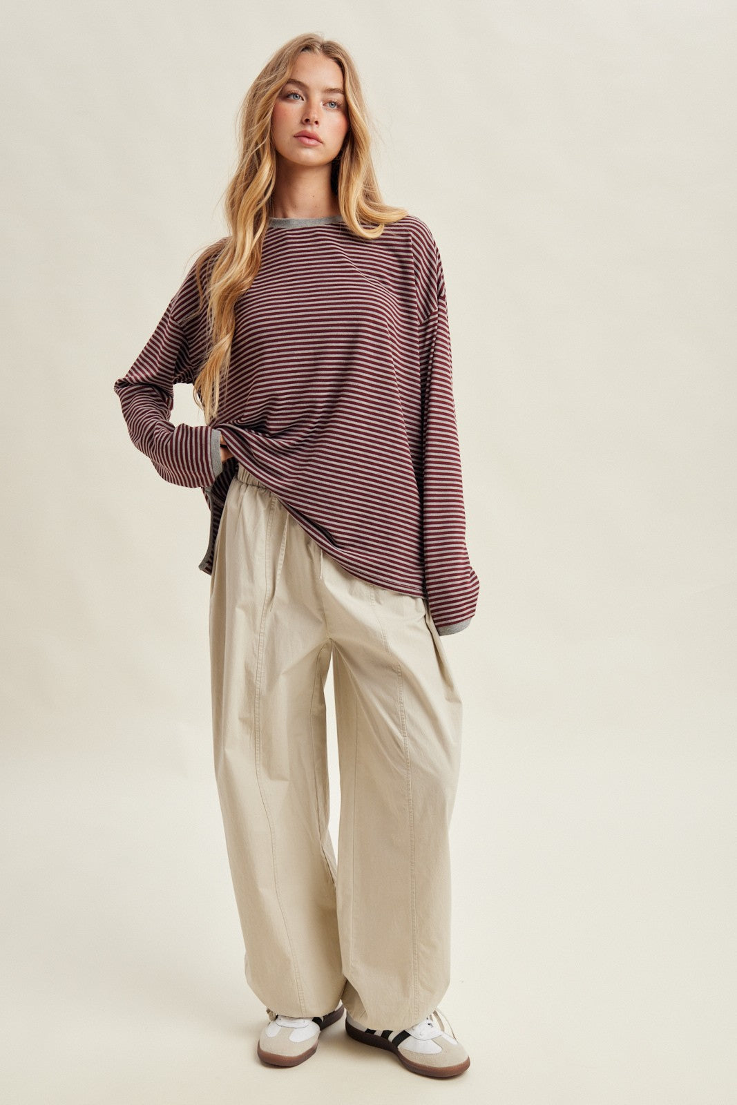woman wearing a maroon and white striped long sleeve top and cream wide-leg pants, standing on a beige background