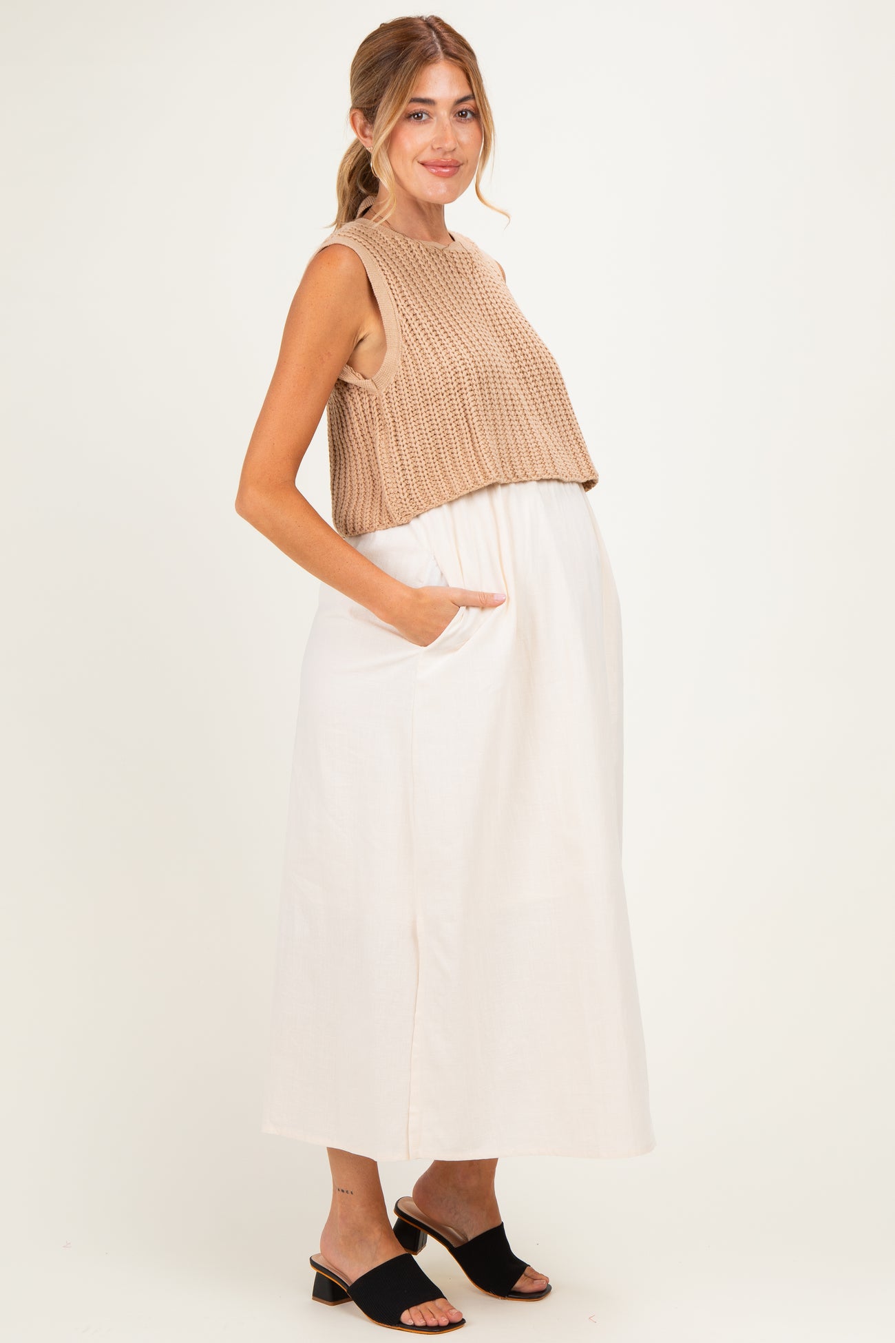 woman wearing a beige knitted crop top and a white long maternity dress with pockets, showing her baby bump