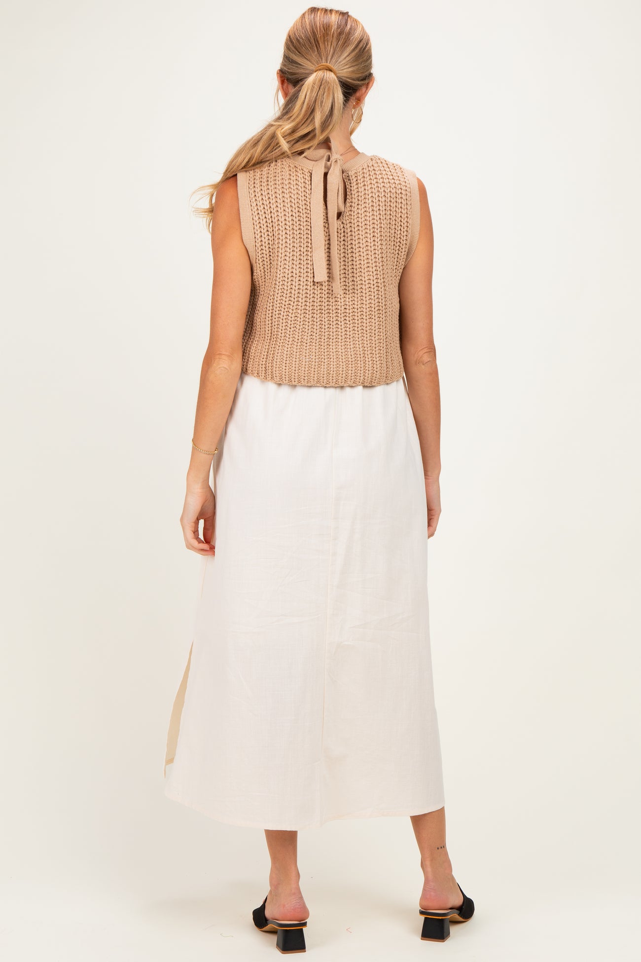 woman wearing a beige knitted maternity vest over a long white linen dress, showing a relaxed style with a back tie detail