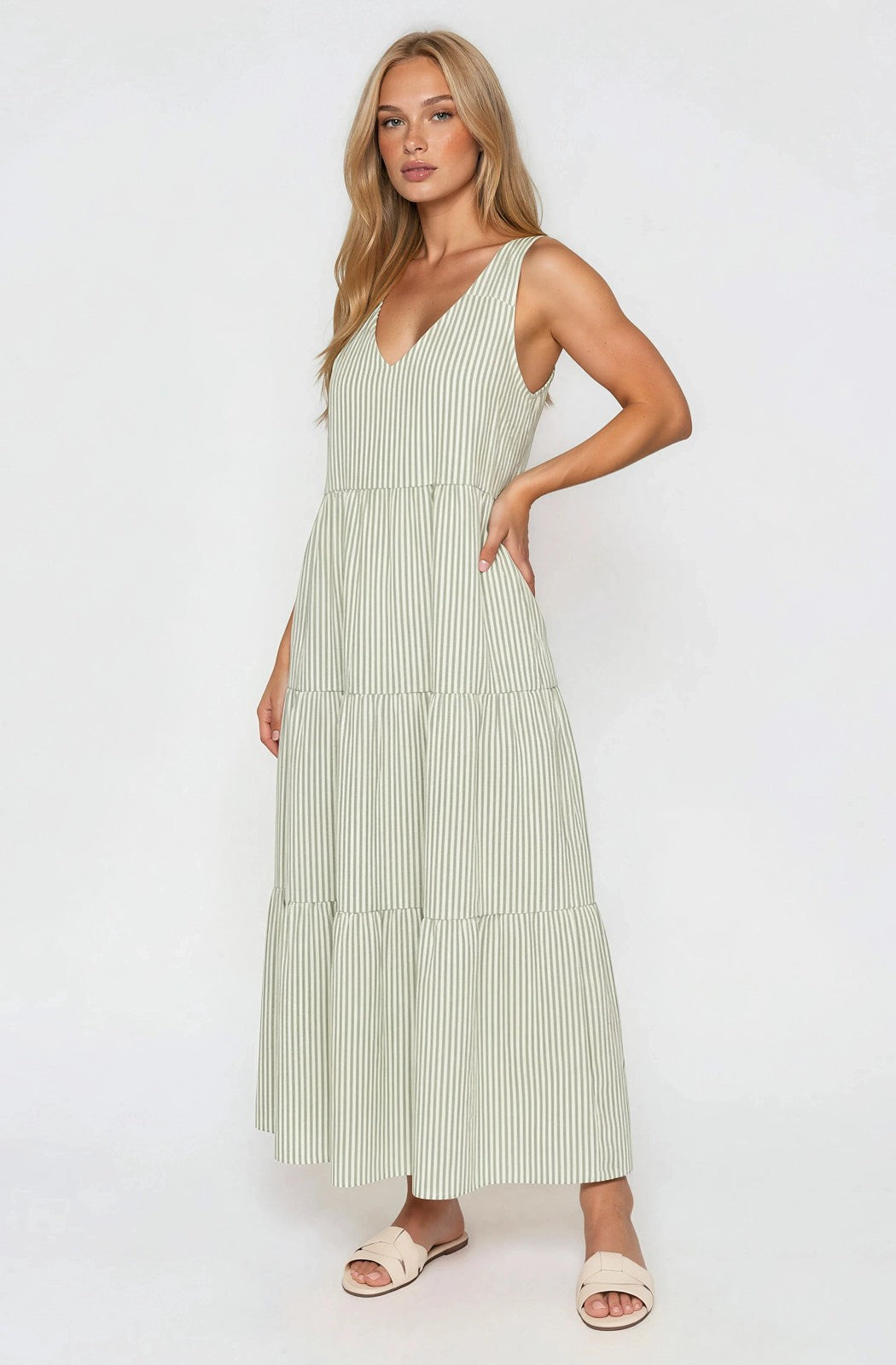 woman wearing a light green striped maxi dress with a v-neck and tiered skirt, standing sideways with hands in pockets