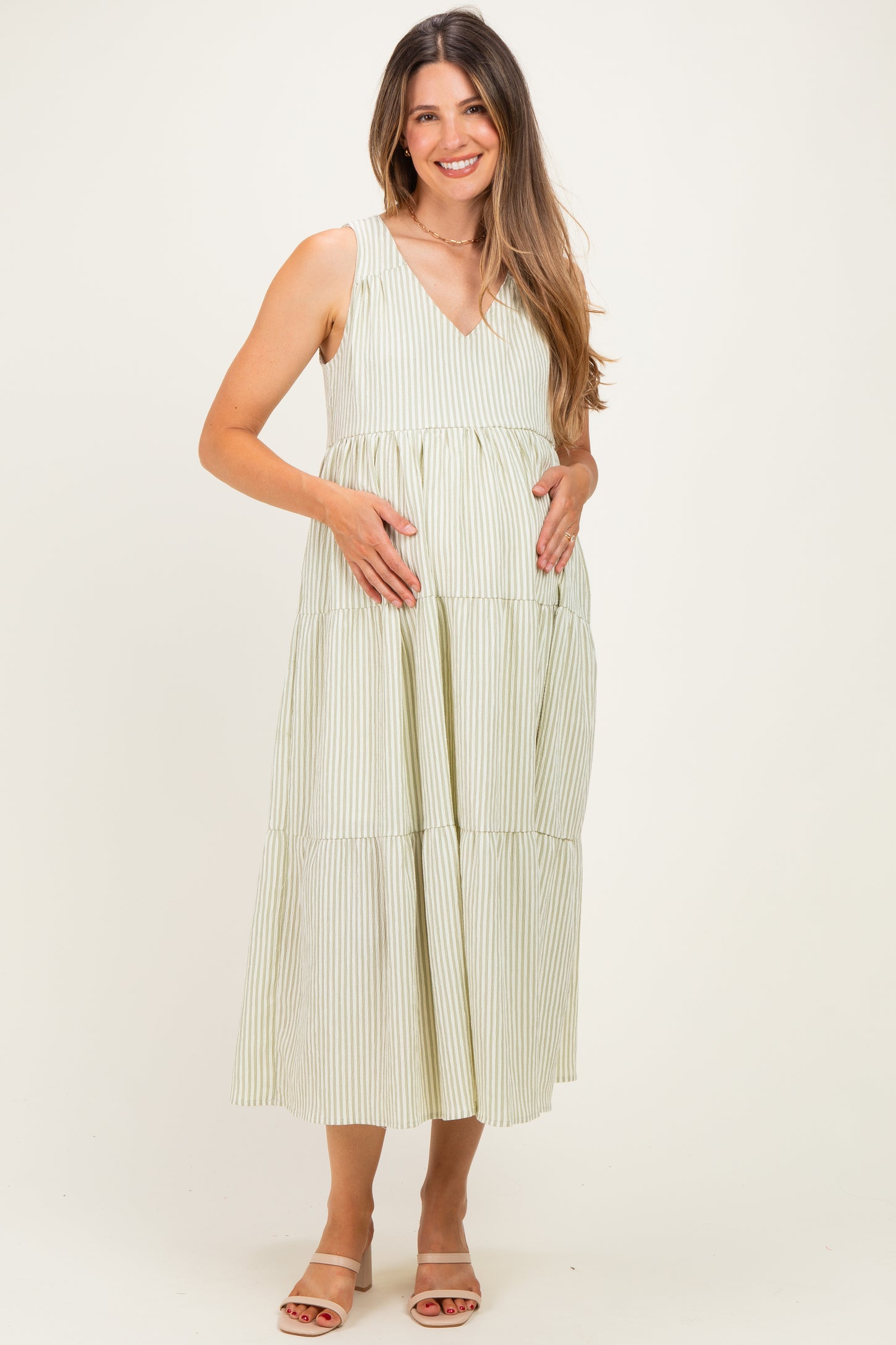 woman wearing a light green and white striped maternity dress, standing and cradling her baby bump
