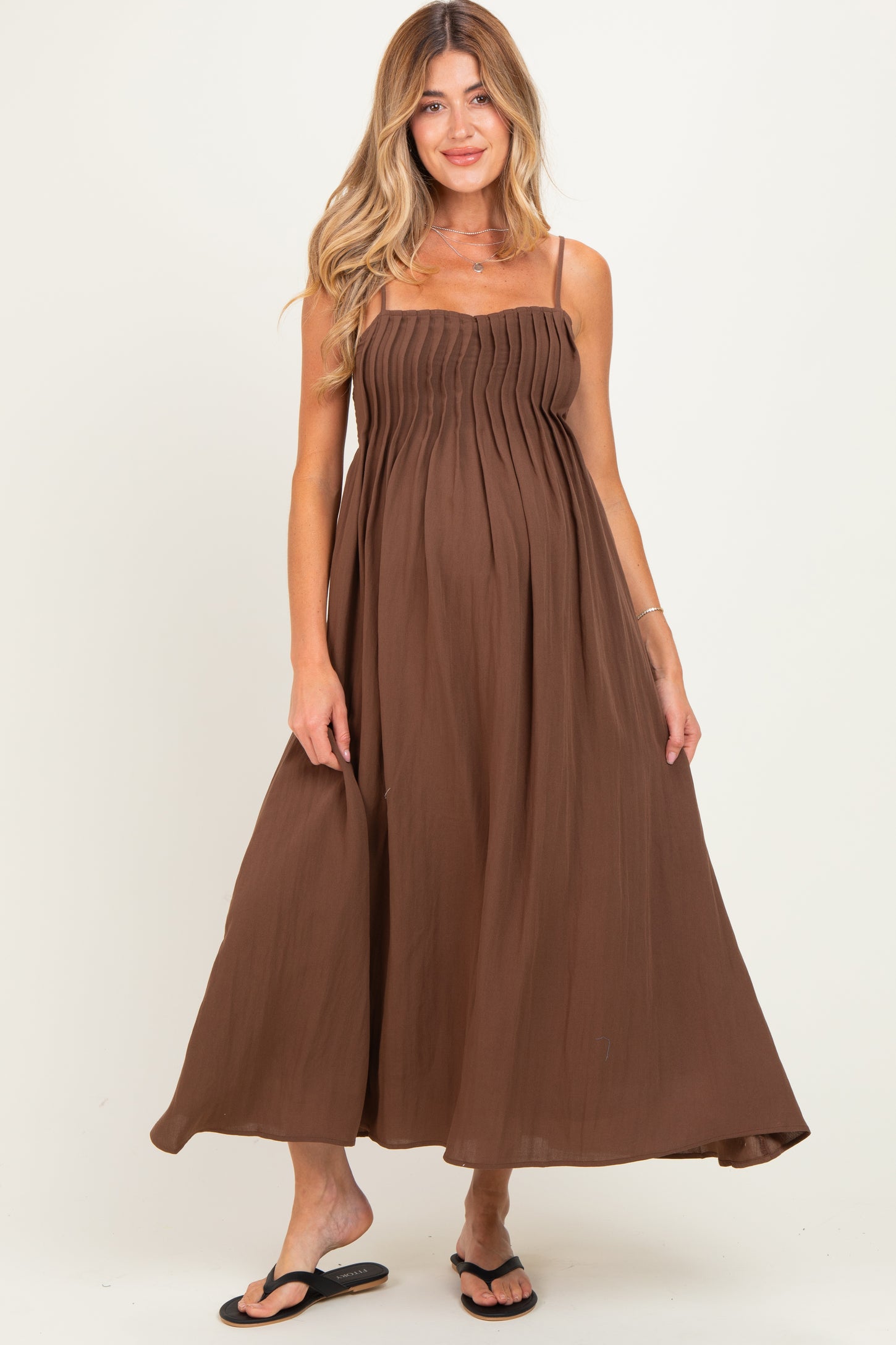 woman wearing a brown maternity maxi dress with adjustable spaghetti straps and a pleated bodice, showing her baby bump