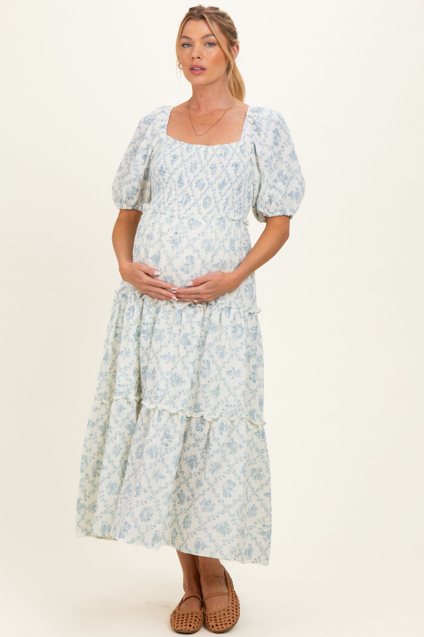 woman wearing a blue patterned maternity dress with puff sleeves and a tiered skirt, cradling her baby bump