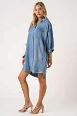 woman wearing a light blue denim shirt dress with a front pocket and rolled sleeves, standing in a relaxed pose