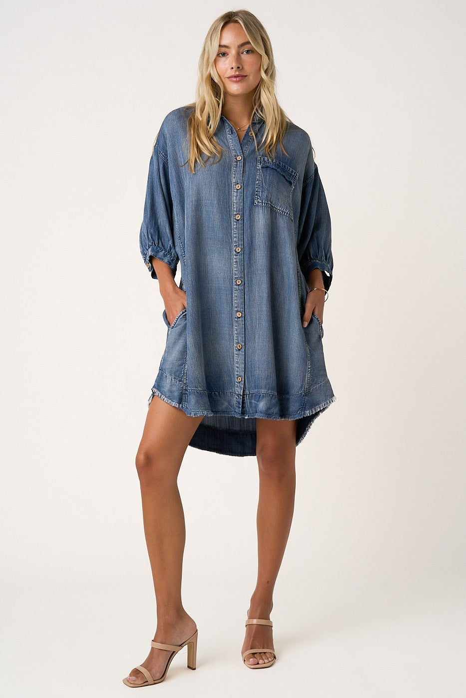 woman wearing a blue denim button-up dress with puff sleeves and side pockets, standing with one hand in her pocket