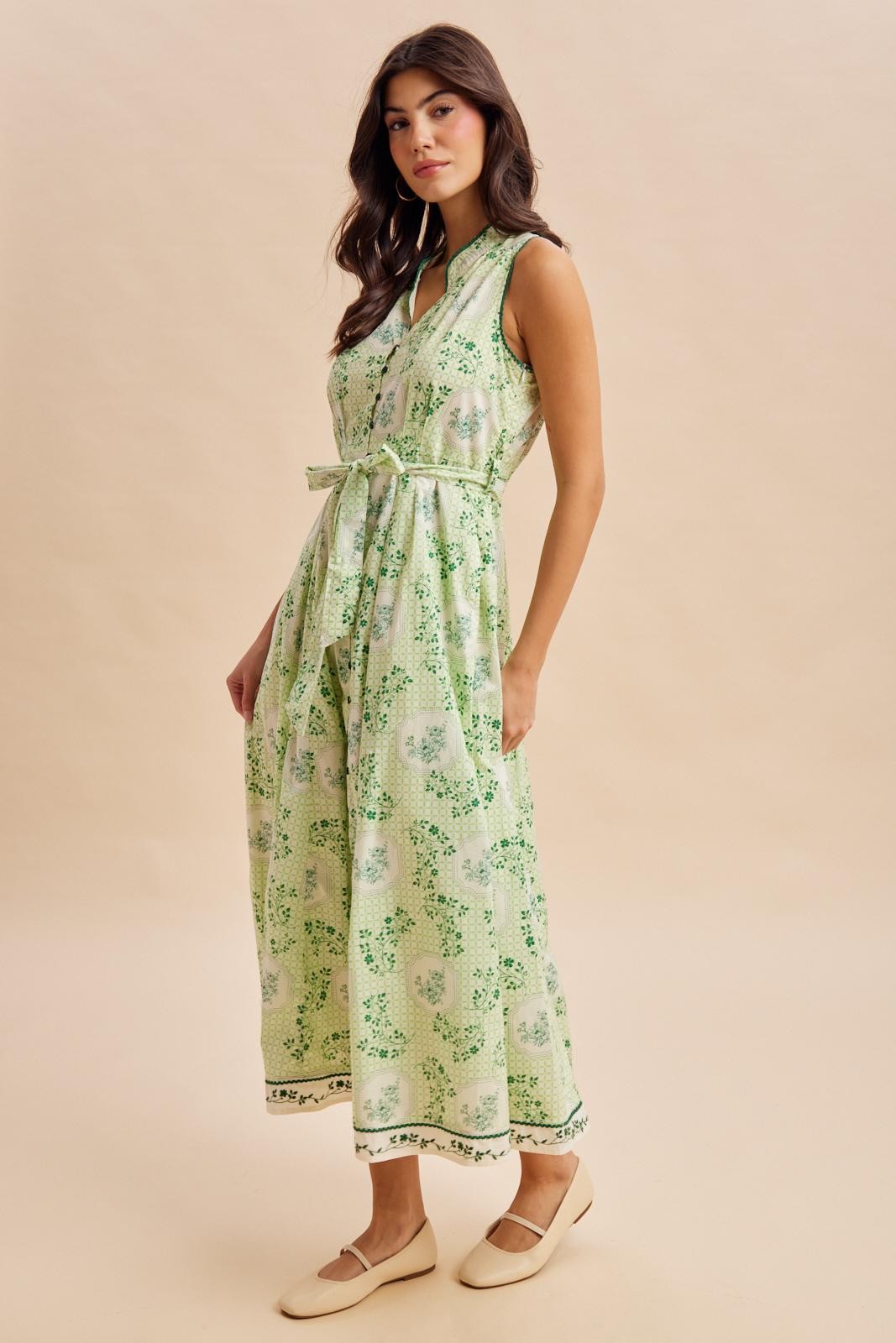 woman wearing a green floral print sleeveless dress with a tie waist detail and flat shoes