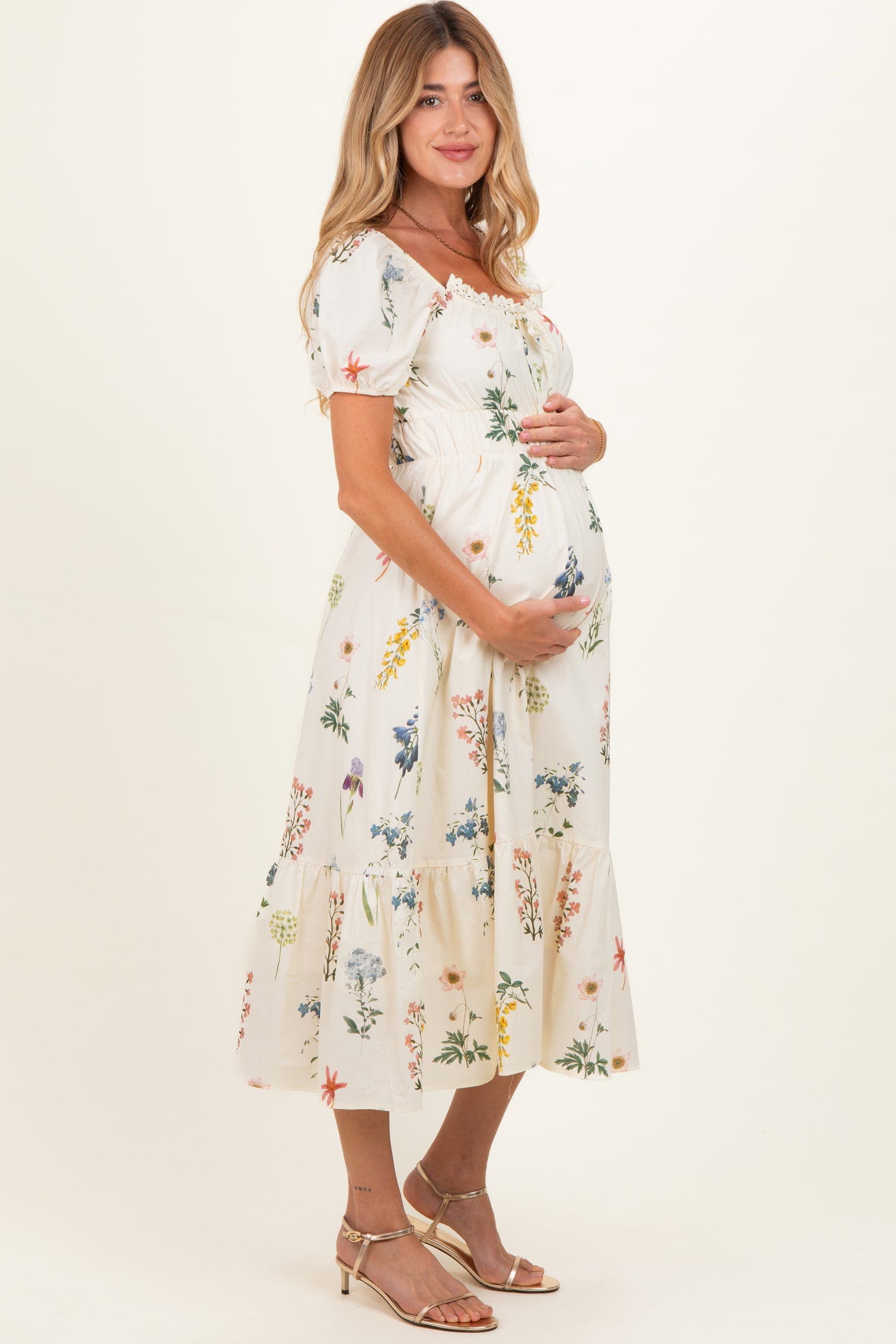 woman wearing a cream floral maternity dress, holding her baby bump with a tiered skirt and short puff sleeves