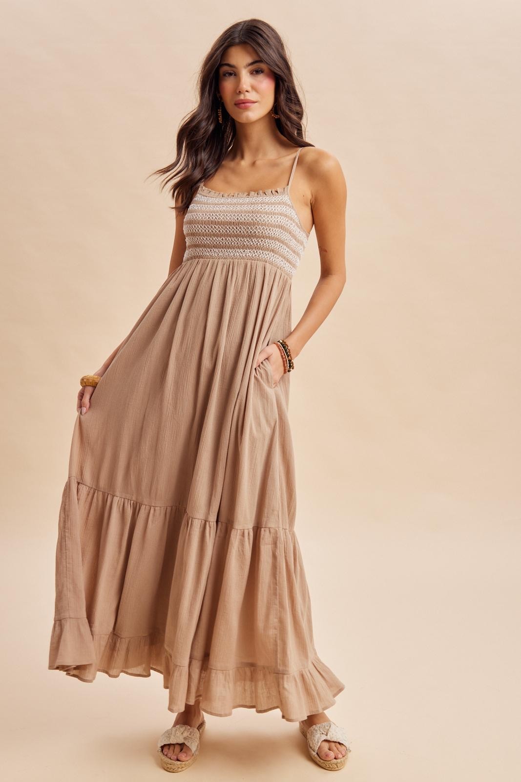 woman wearing a tan maxi dress with spaghetti straps, smocked bodice, and tiered skirt, standing with hands in pockets