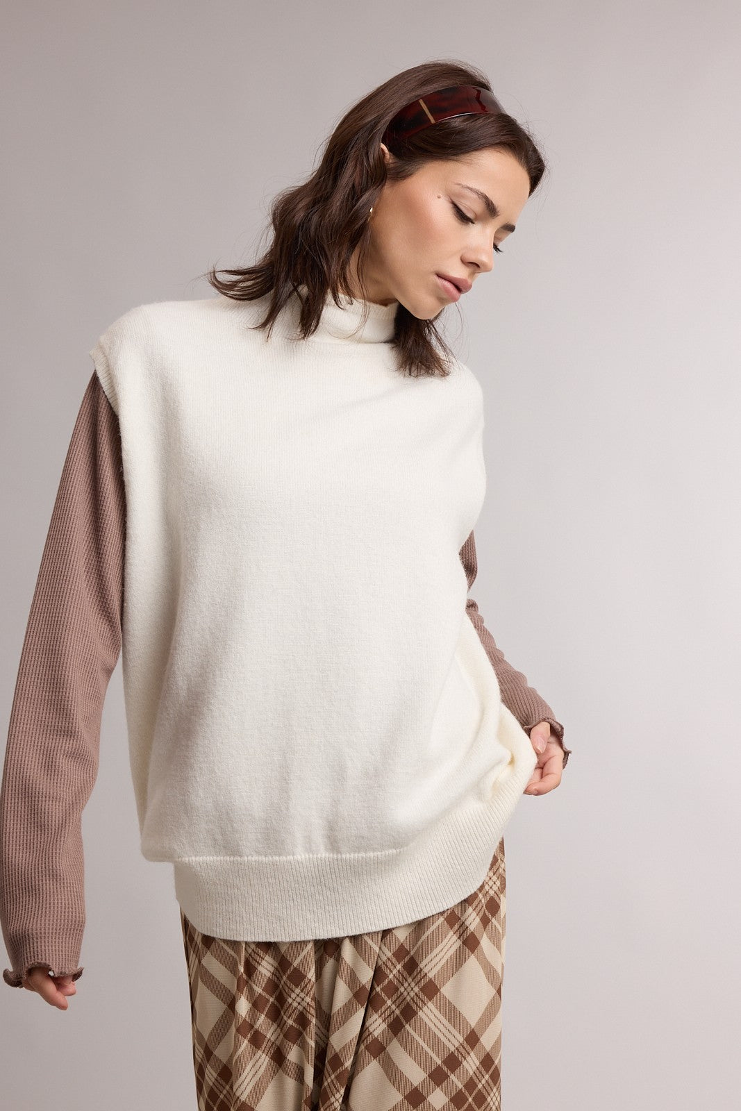 woman wearing a white sleeveless turtleneck sweater over a brown long sleeve top and plaid wide-leg trousers