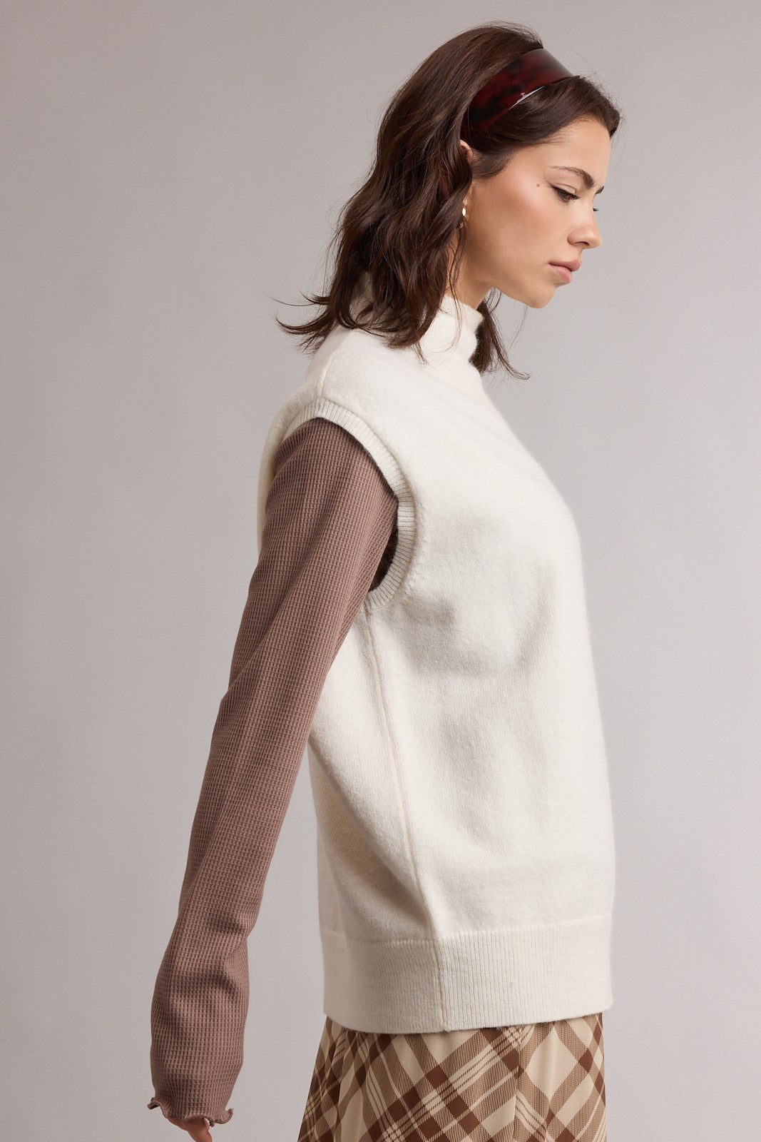 woman wearing a white sleeveless turtleneck sweater over a brown long sleeve top and a plaid skirt