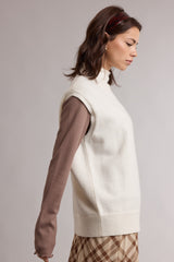 woman wearing a white sleeveless turtleneck sweater over a brown long sleeve top and a plaid skirt