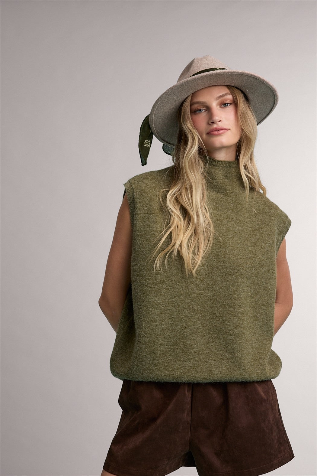 woman wearing an olive green sleeveless sweater with a high neck and brown suede shorts, styled with a hat and scarf