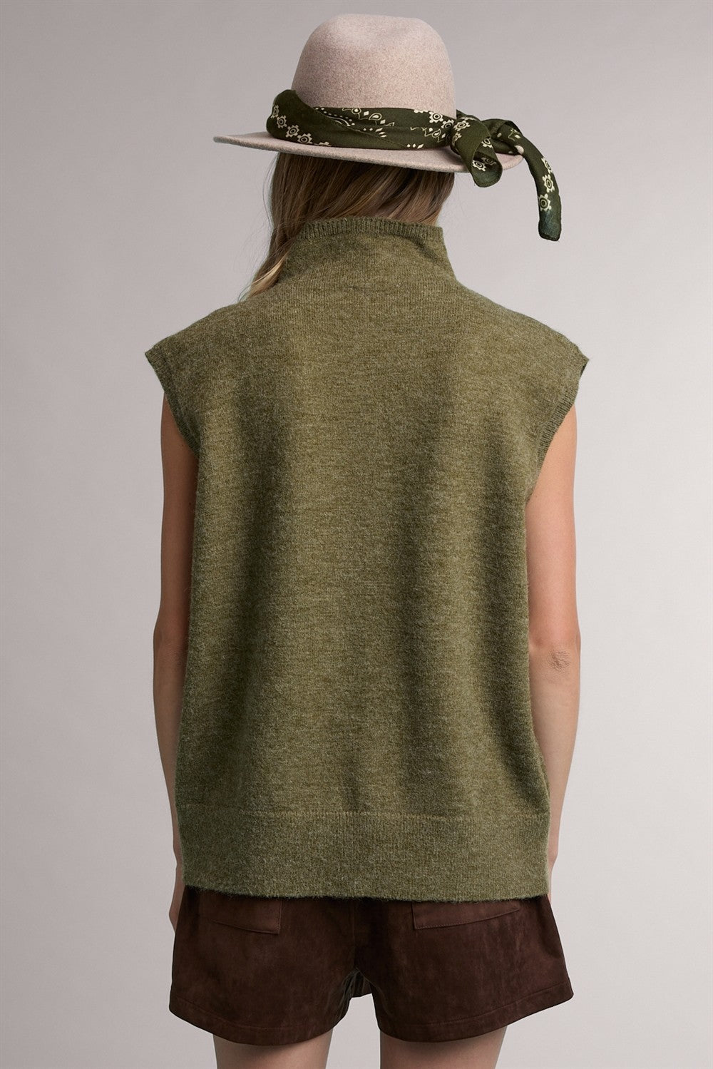 woman wearing an olive green sleeveless sweater with a high neckline, accessorized with a grey hat and green scarf