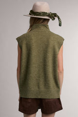 woman wearing an olive green sleeveless sweater with a high neckline, accessorized with a grey hat and green scarf