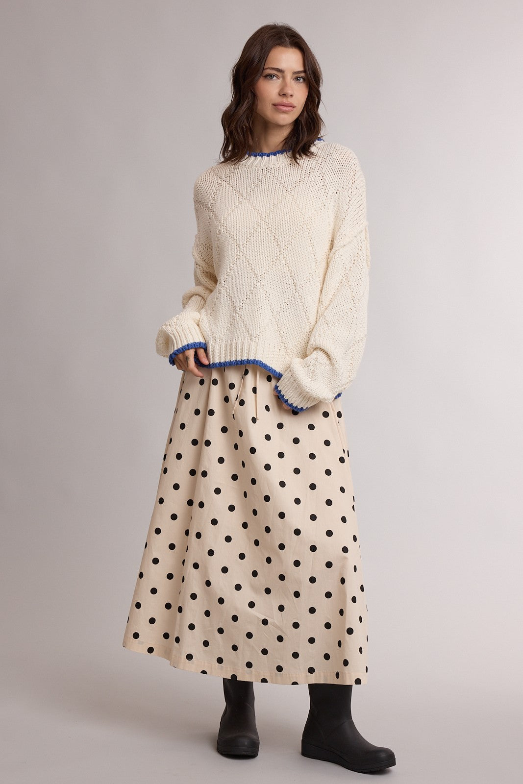 woman wearing a cream sweater with blue trim and a beige polka dot maxi skirt, paired with black ankle boots