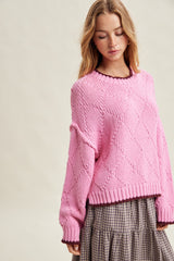 woman wearing a pink knitted sweater with a diamond pattern and a black-trimmed neckline over a plaid skirt