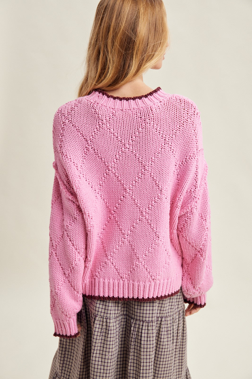 woman wearing a pink oversized knit sweater with a diamond pattern, shown from the back over a plaid skirt
